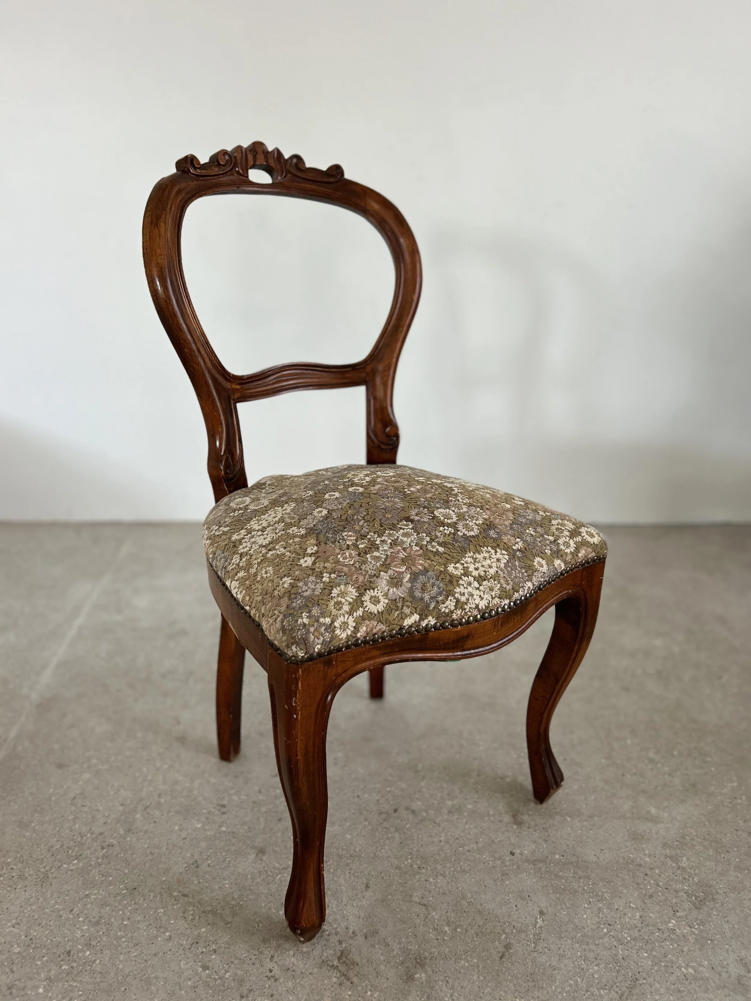 An antique wooden chair with an ornate backrest and a cushioned seat covered in floral upholstery, placed against a plain light-colored background.