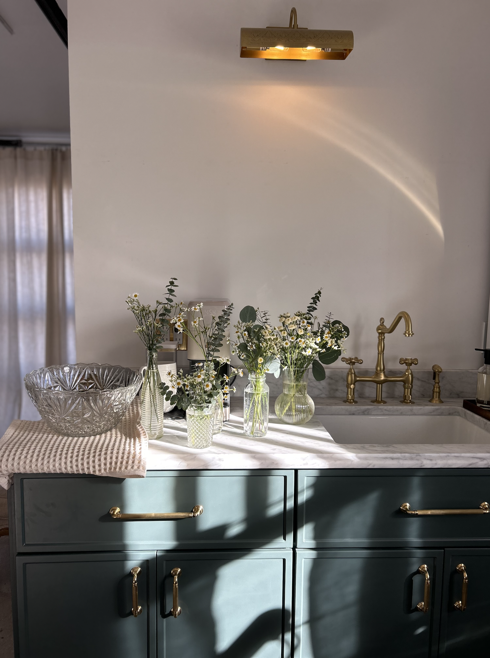 Kitchen counter with green cabinetry, gold hardware, marble countertop, a sink with a gold faucet, and three vases with white and green flowers, illuminated by a gold wall-mounted light.