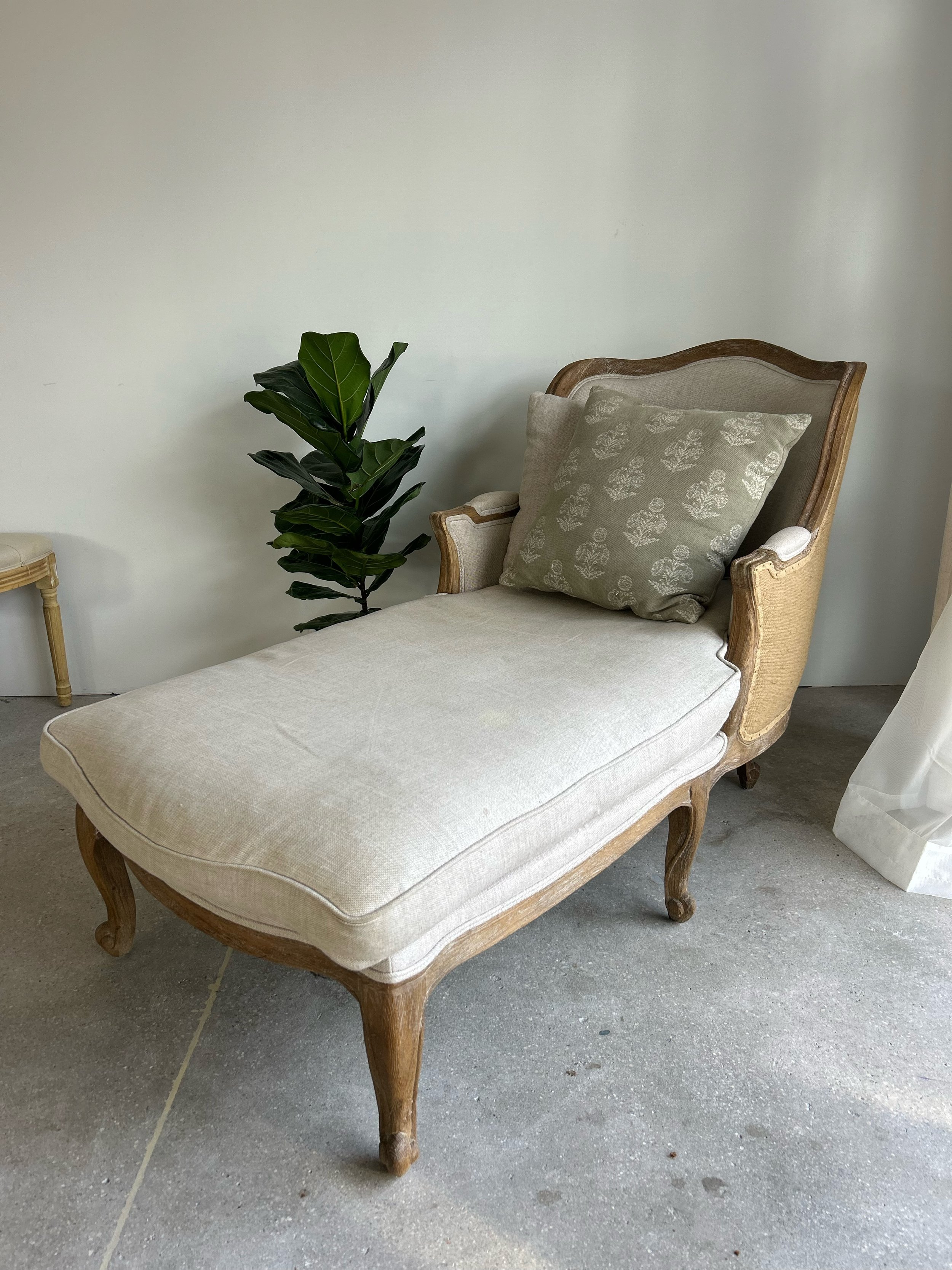 A vintage beige chaise lounge with wooden frame, a decorative pillow, positioned next to a green leafy plant.
