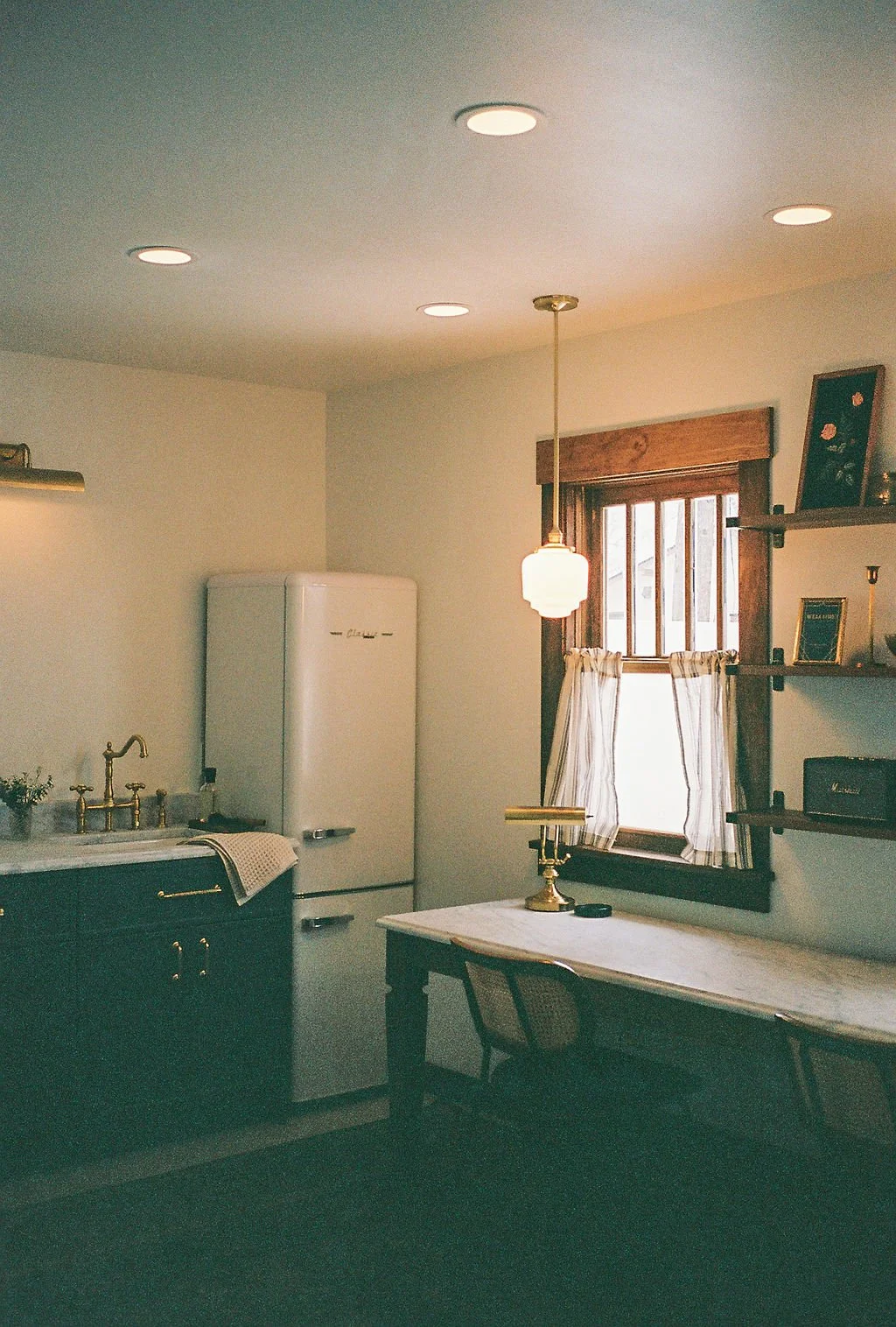 Vintage kitchen with white refrigerator, black cabinets, marble countertop, gold faucet, small potted plant, hanging pendant light, wooden window with white curtains, wooden shelves with photos and decor, and a marble-top dining table with a brass ca