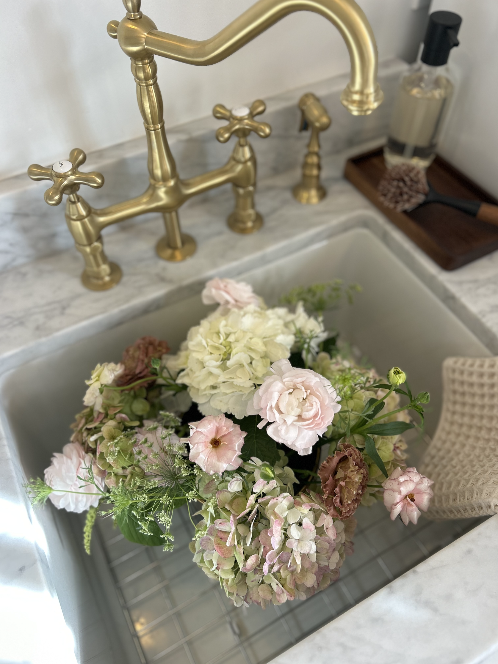 A bouquet of light pink, white, and green flowers in a kitchen sink with a gold faucet.