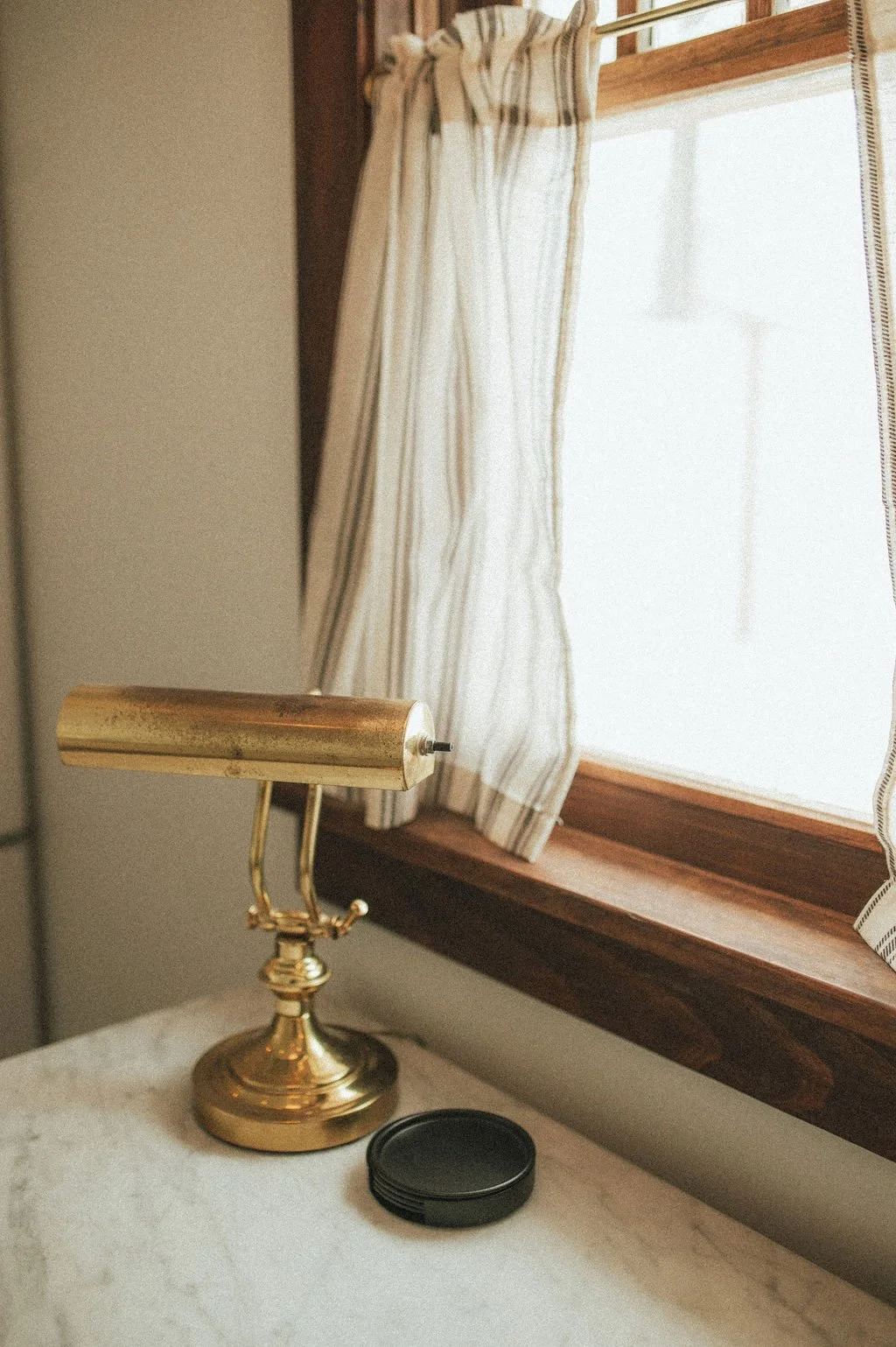 A gold desk lamp, a black coaster, and a window with white curtains and wooden trim.