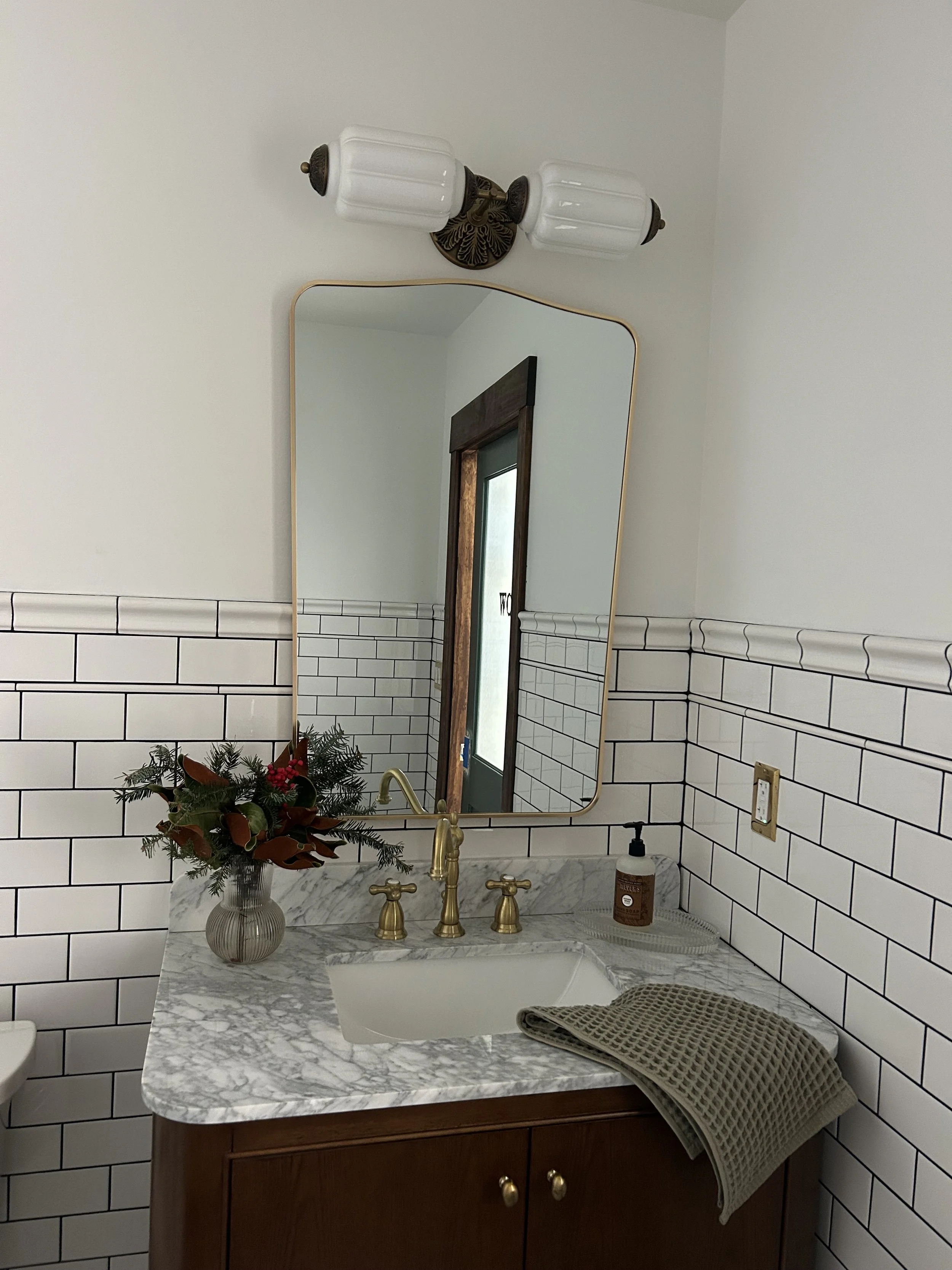 Bathroom vanity with a marble countertop, an oval sink, gold faucet, a mirror, wall-mounted light fixtures, a vase with holiday flowers, a hand soap dispenser, and a towel.