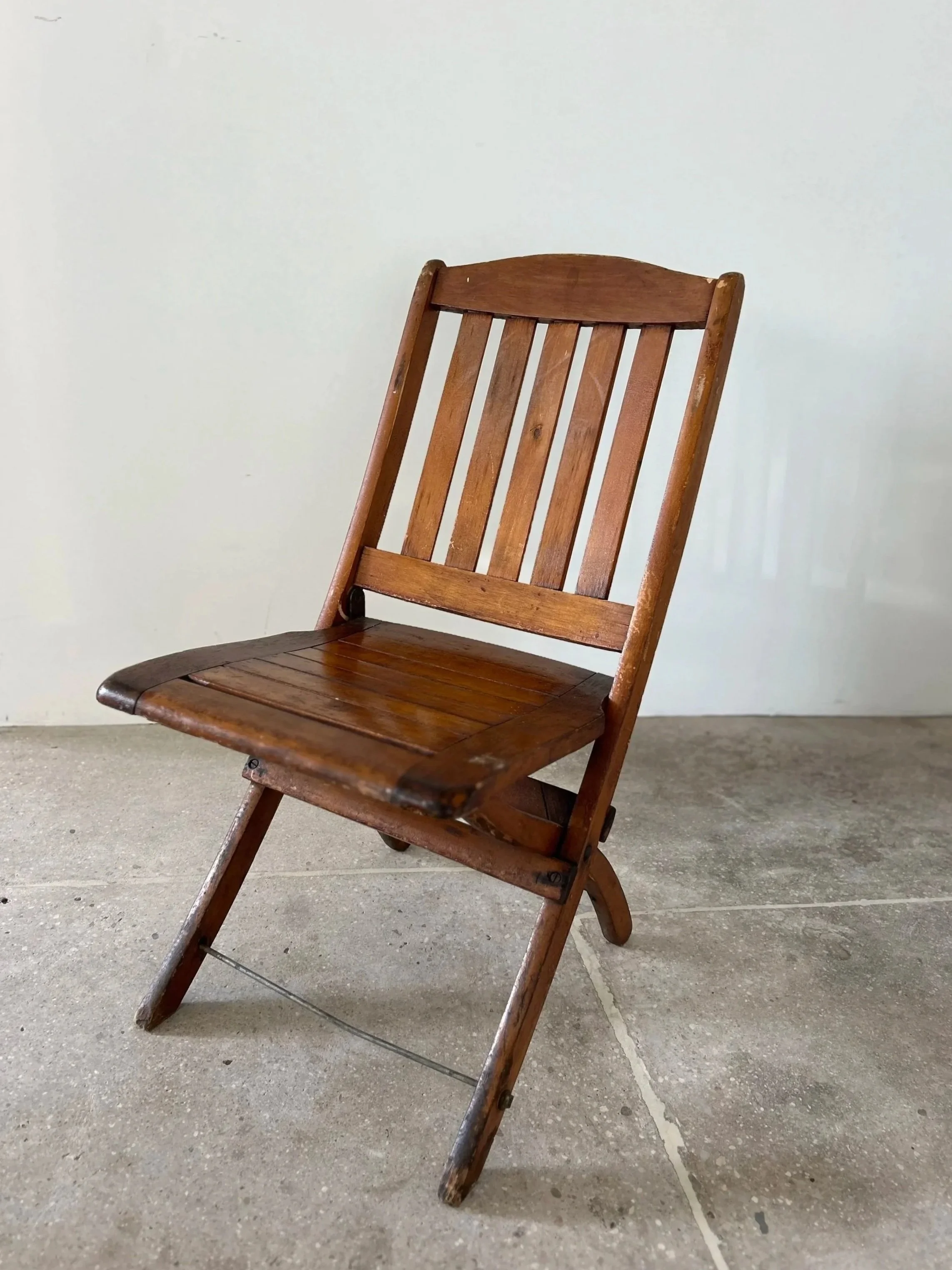 A wooden folding chair with a slatted backrest and seat, placed on a concrete floor against a plain white wall.