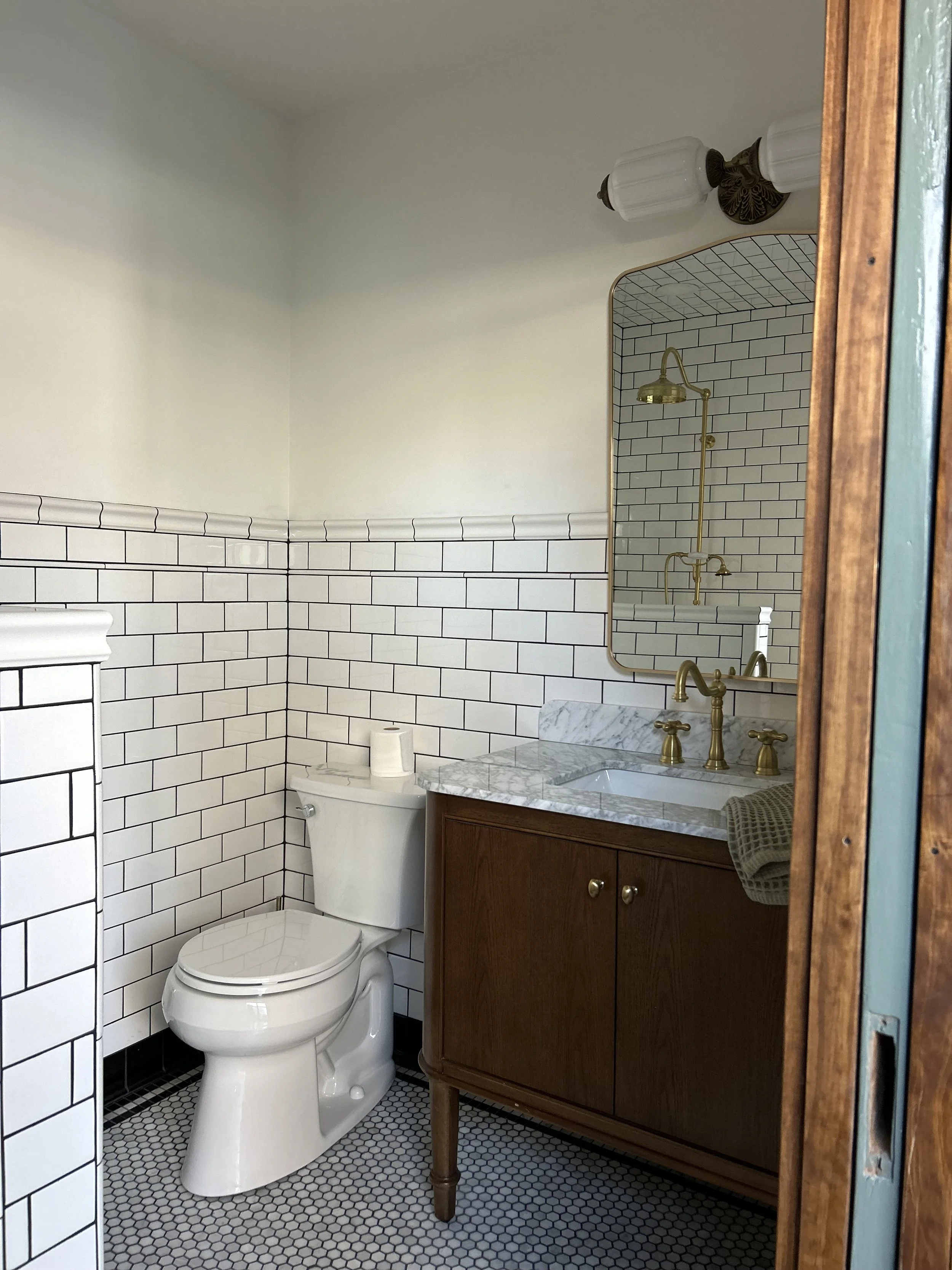 A bathroom with white subway tiles on the lower half of the wall, a white toilet, a wooden vanity with a marble countertop, and a mirror above the sink. There is a brass faucet and fixtures, and a wall-mounted light fixture with two white glass shade