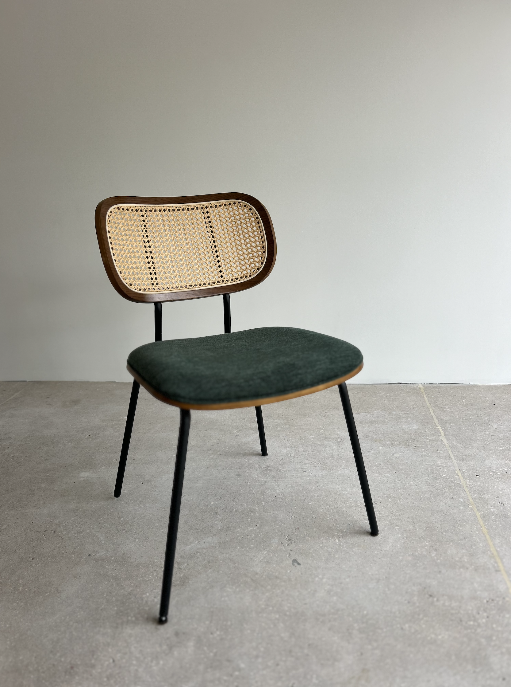 A vintage chair with black metal legs, a woven cane backrest, and a dark green upholstered seat, placed against a plain white wall.