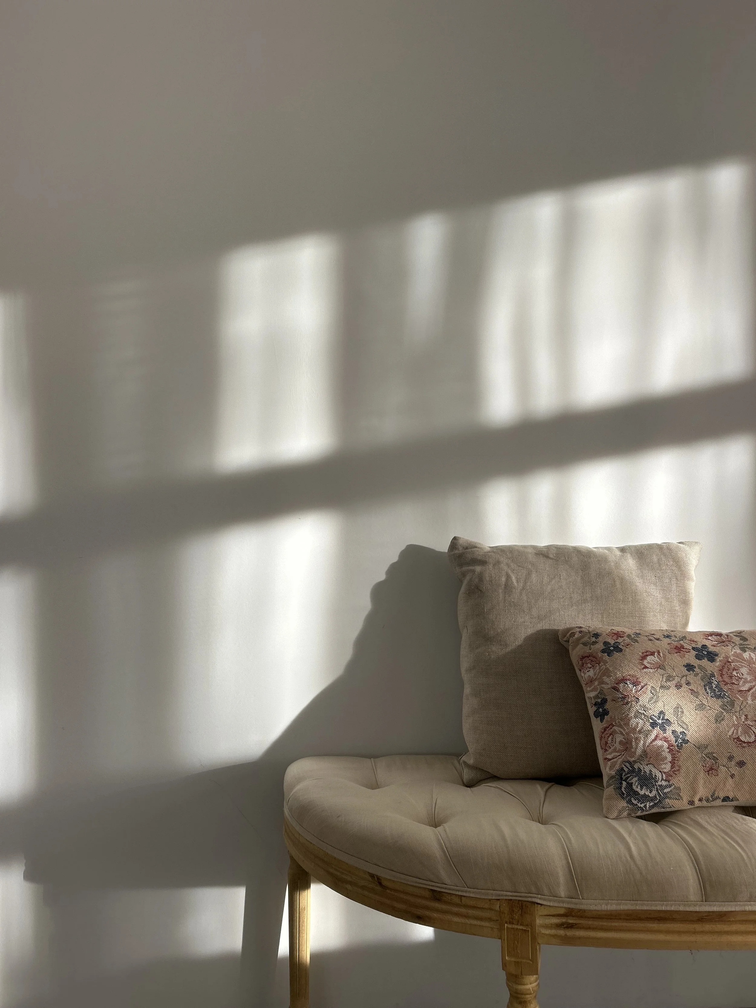 Sunlight casting shadows of window panes on a white wall, with a beige cushioned bench topped with two pillows, one beige and one floral patterned.