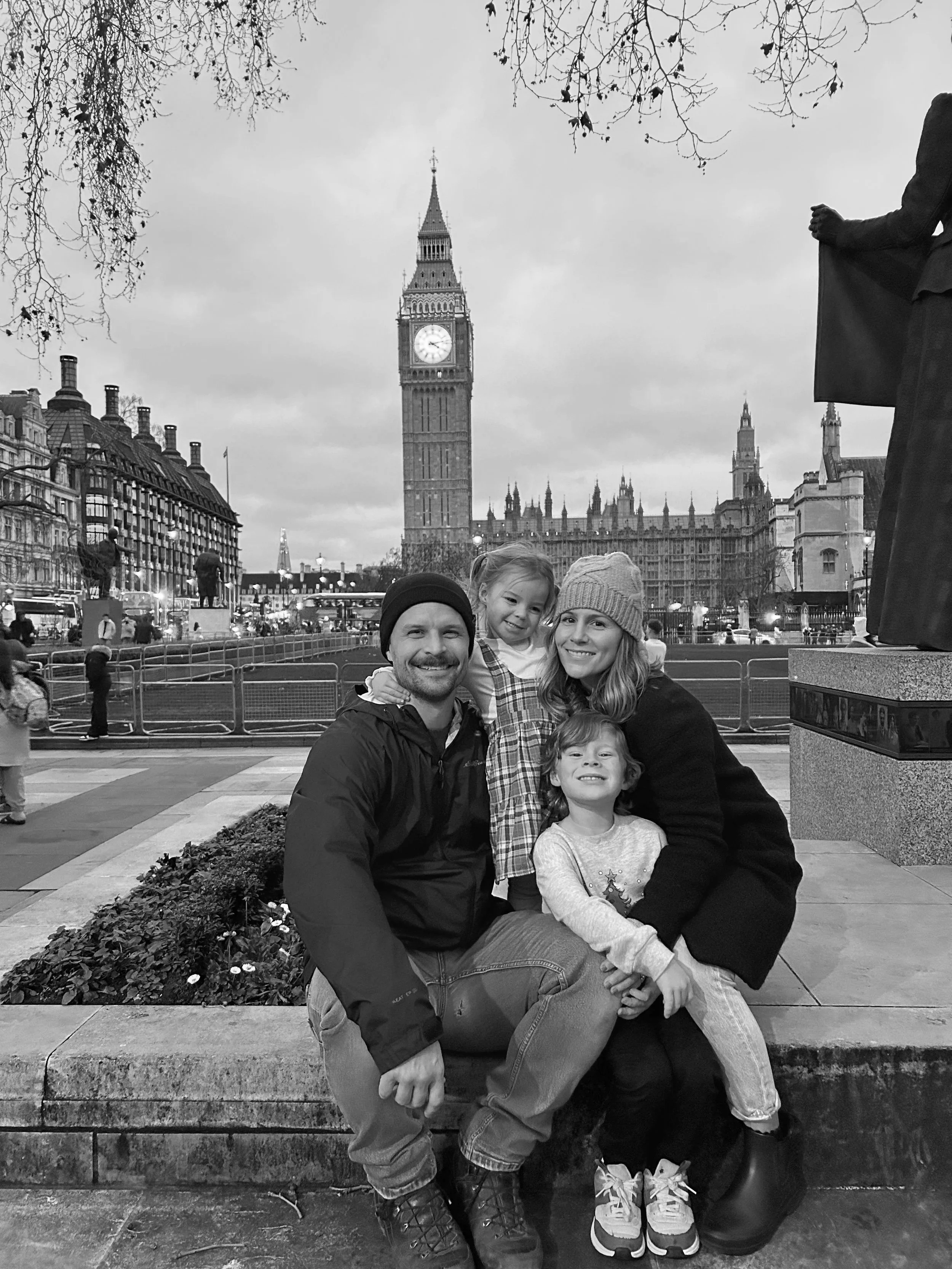 A family of four, two adults and two children, smiling and sitting on a low wall in front of Big Ben and the Houses of Parliament in London. The image is in black and white.