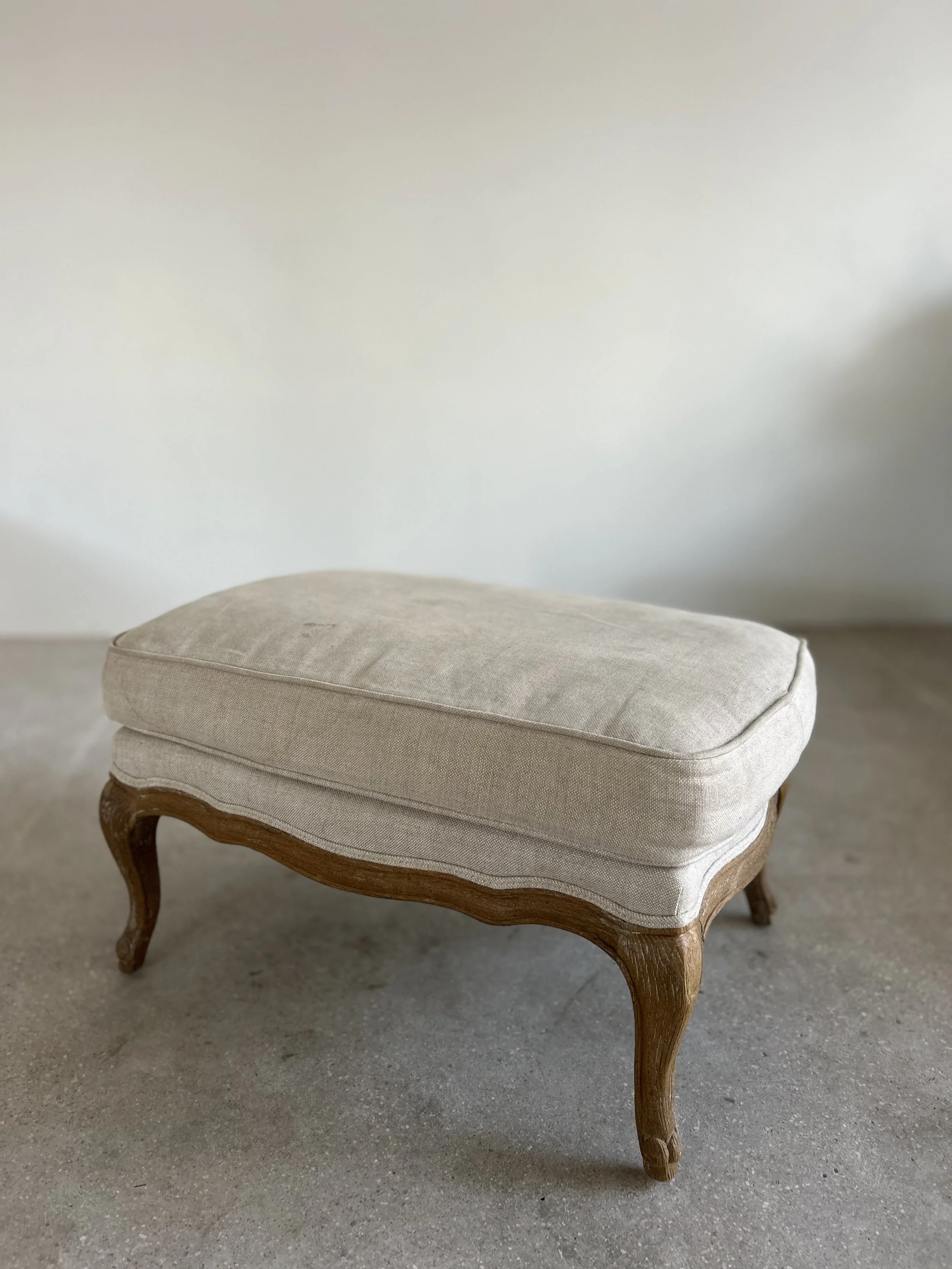 A beige upholstered ottoman with wooden carved legs on a concrete floor against a plain white wall.