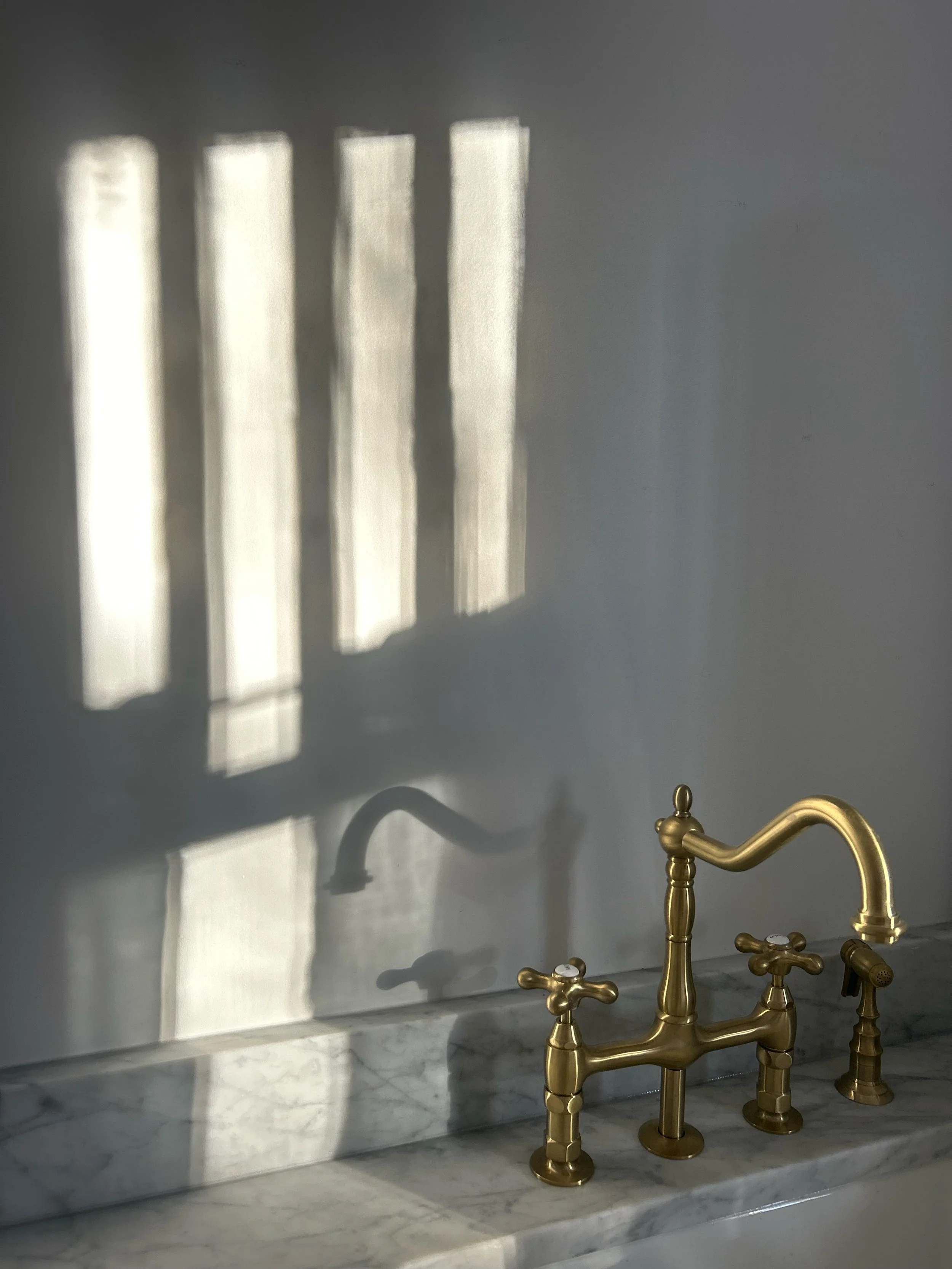 Brass kitchen faucet with two handles mounted on a marble countertop, with sunlight casting shadows of a window grille on a white wall behind it.