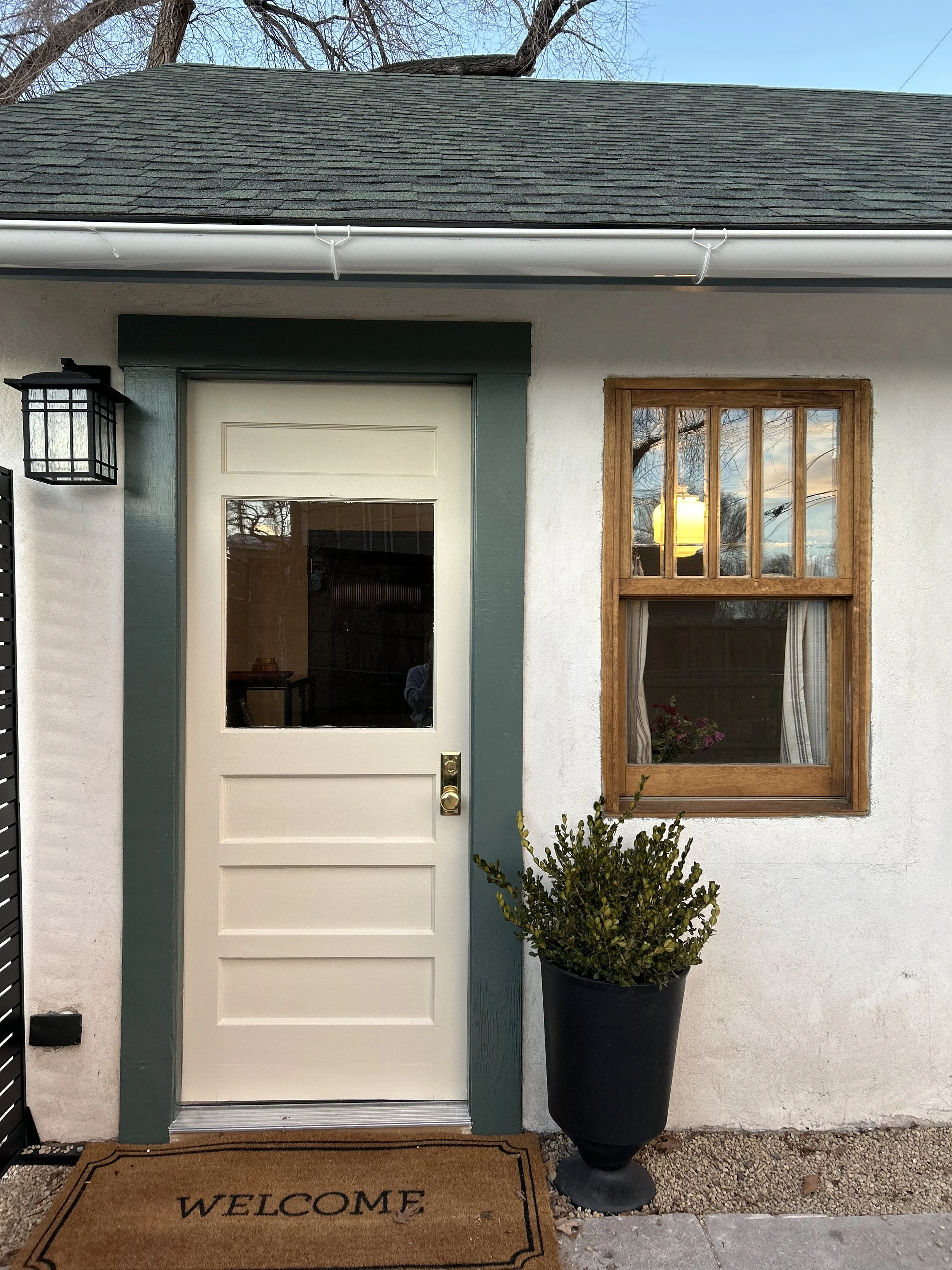 Front entrance of a house with a white door, green trim, a window with curtains and a potted plant outside, and a welcome mat on the ground.