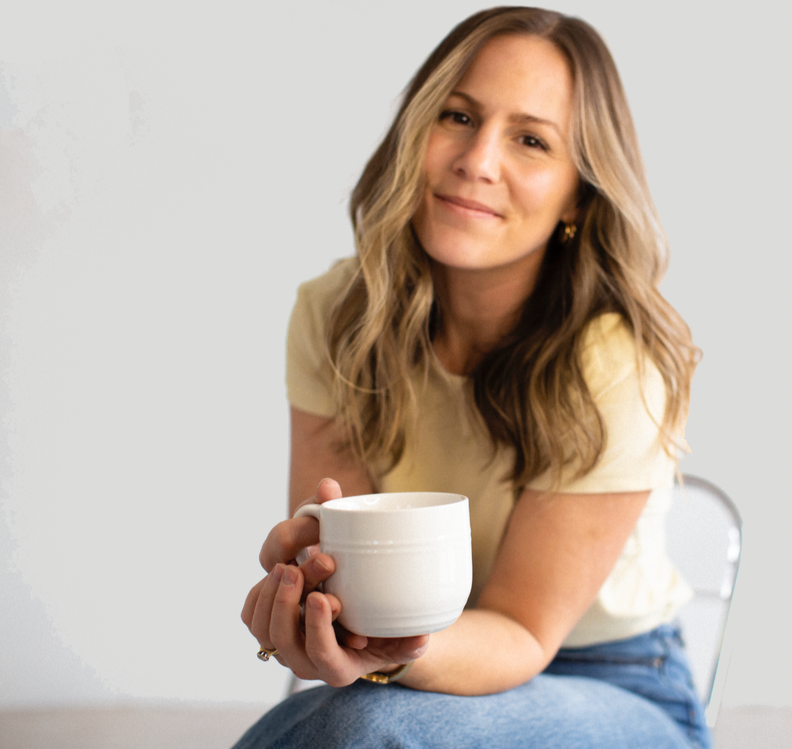 A woman with wavy, shoulder-length blonde hair, wearing a light yellow T-shirt, holding a white ceramic mug and smiling softly while sitting on a chair against a light-colored background.