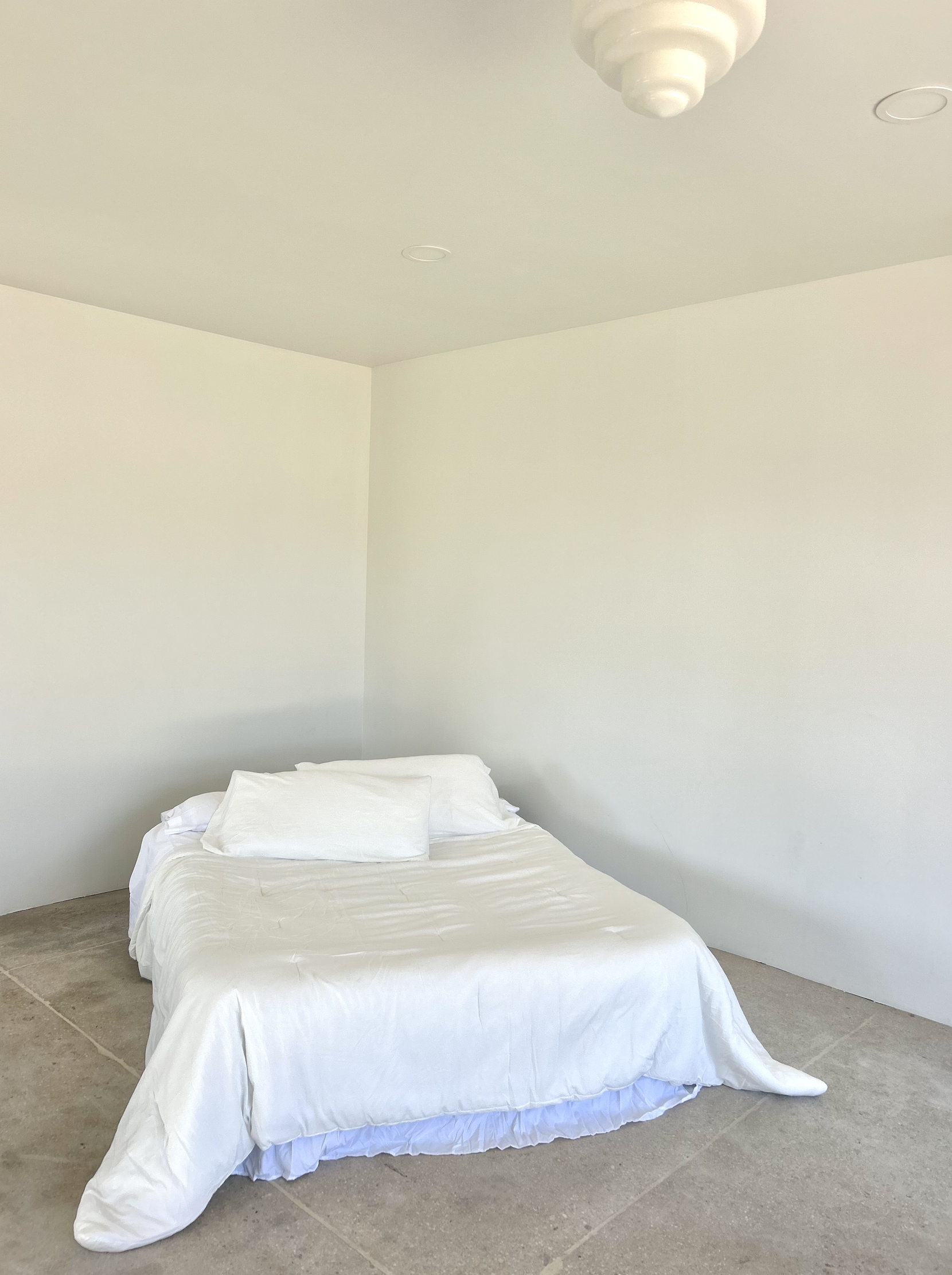 A bed with white bedding and pillows in a room with plain white walls and concrete floor.