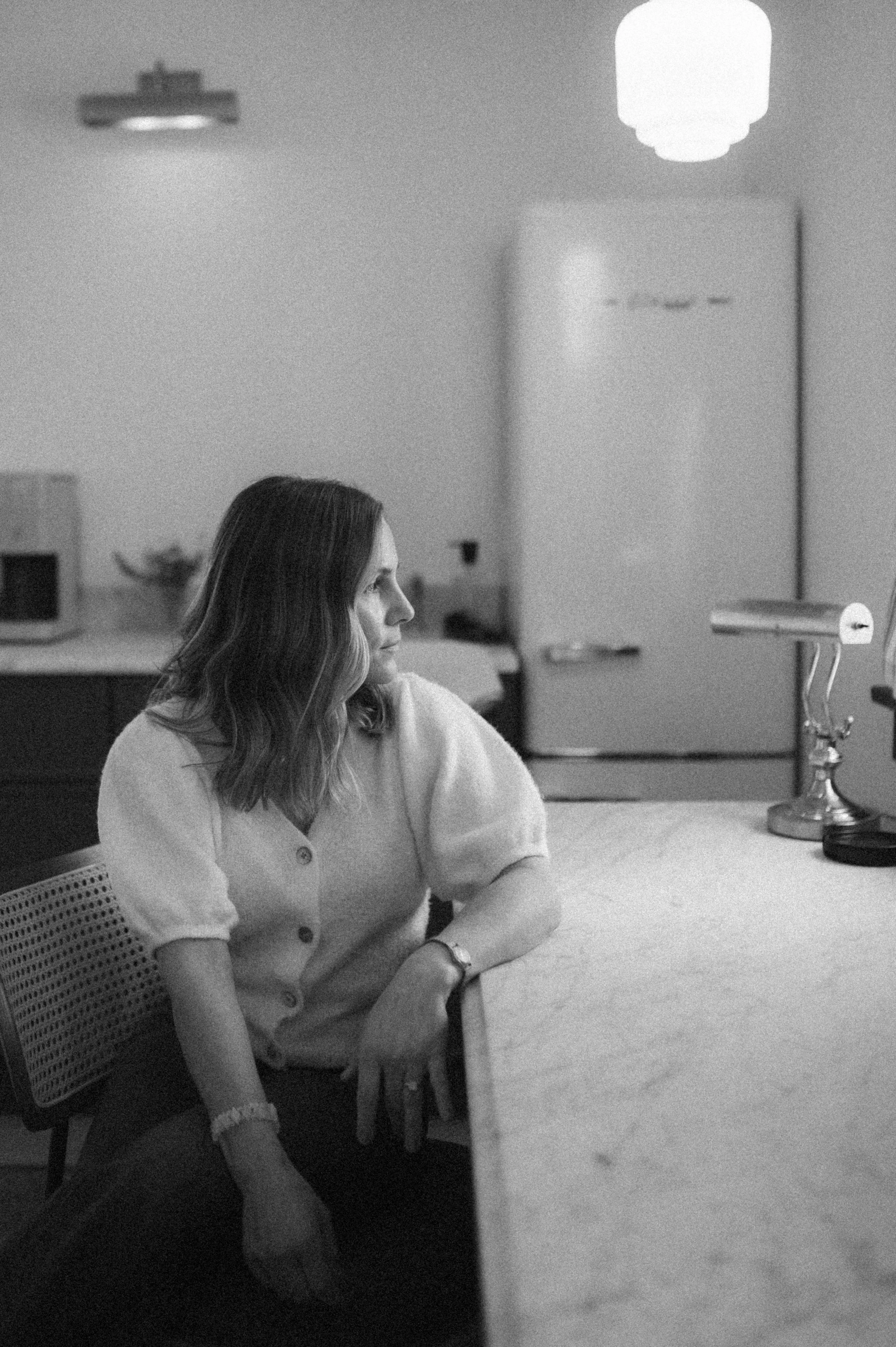 A woman with shoulder-length hair, sitting at a table in a kitchen, looking to her left, wearing a short-sleeved button-up top.