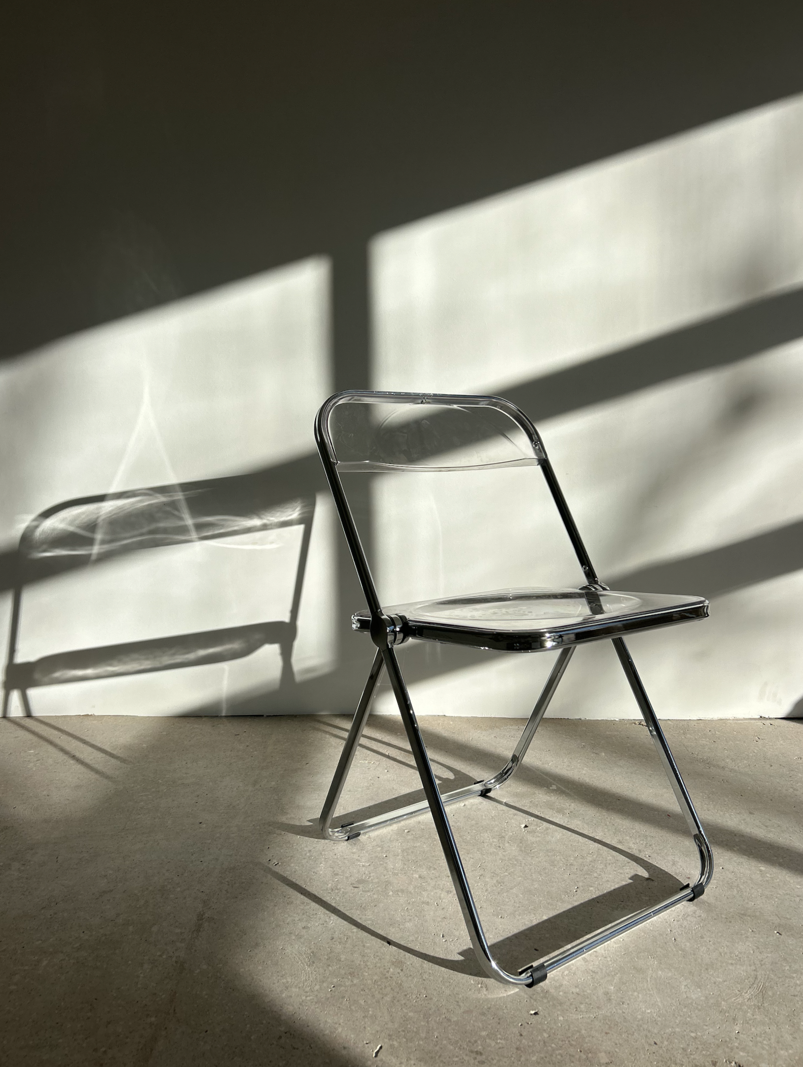 A clear acrylic chair casting a shadow on a white wall, with sunlight creating diagonal patterns.