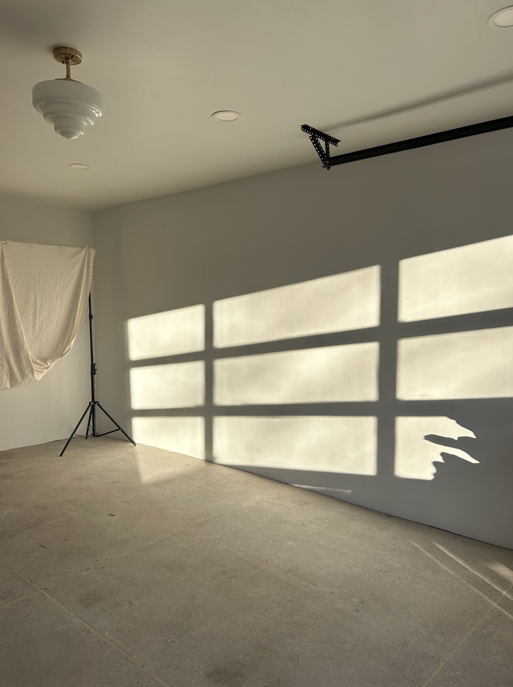 Empty room with beige carpet, a softbox light on a stand, and shadows on the wall cast by a window with sunlight streaming through.