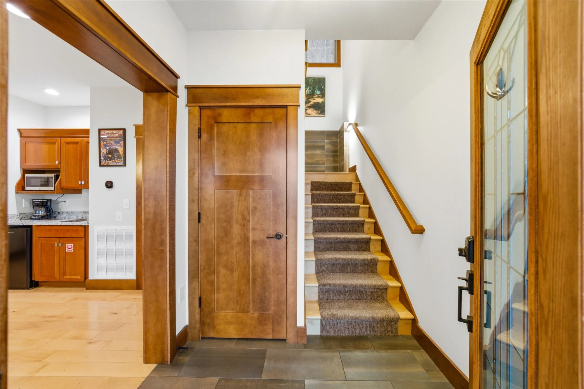 Interior view of a house featuring a wooden staircase with a brown carpet runner, a wooden door in the center, a staircase with a half wall and a window at the top, and a kitchen area with wooden cabinets, a microwave, and a coffee maker.