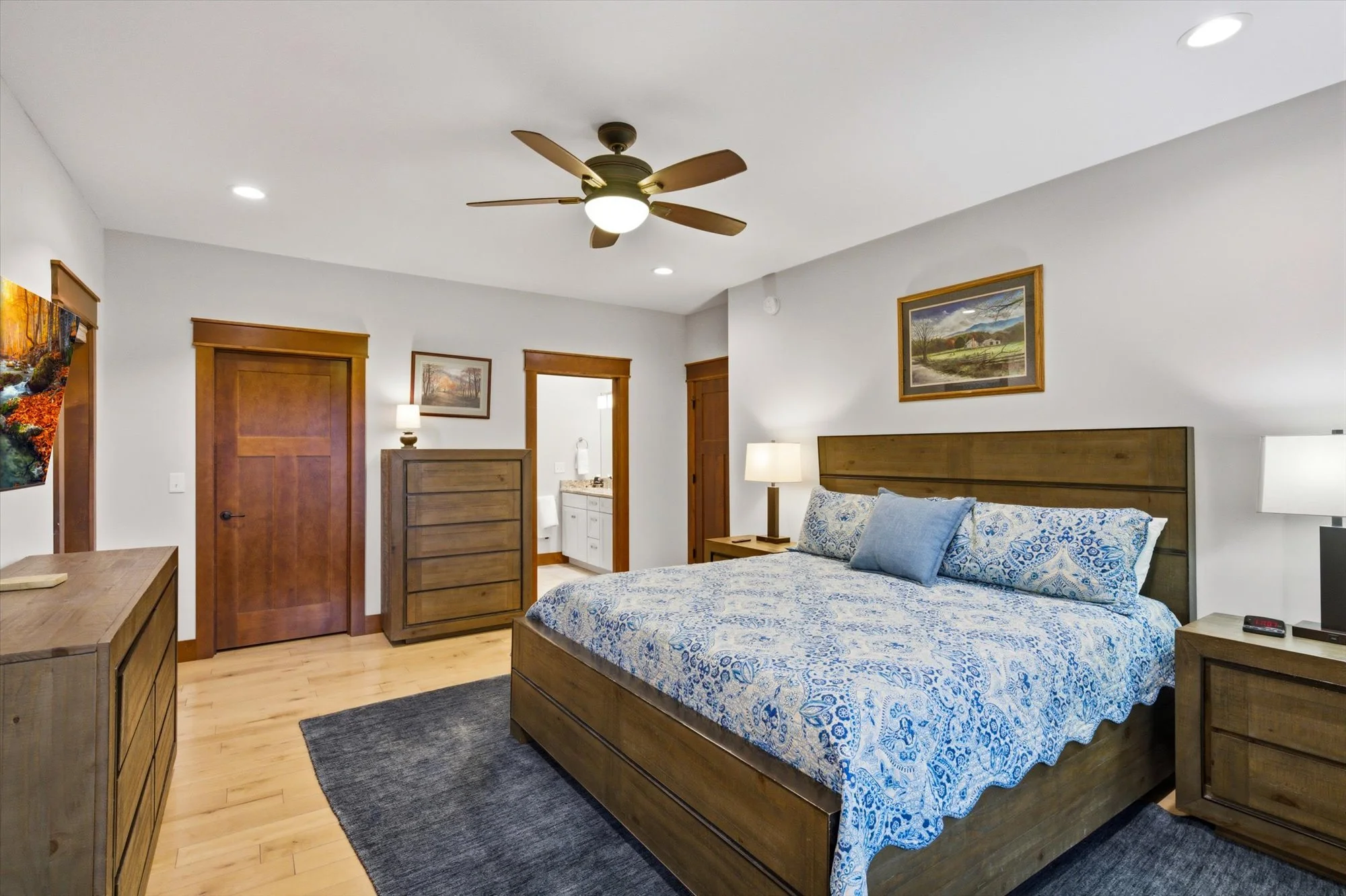 A bedroom with a wooden bed, blue patterned bedding, two bedside tables with lamps, a dresser, a ceiling fan, and two framed landscape paintings on the white walls.