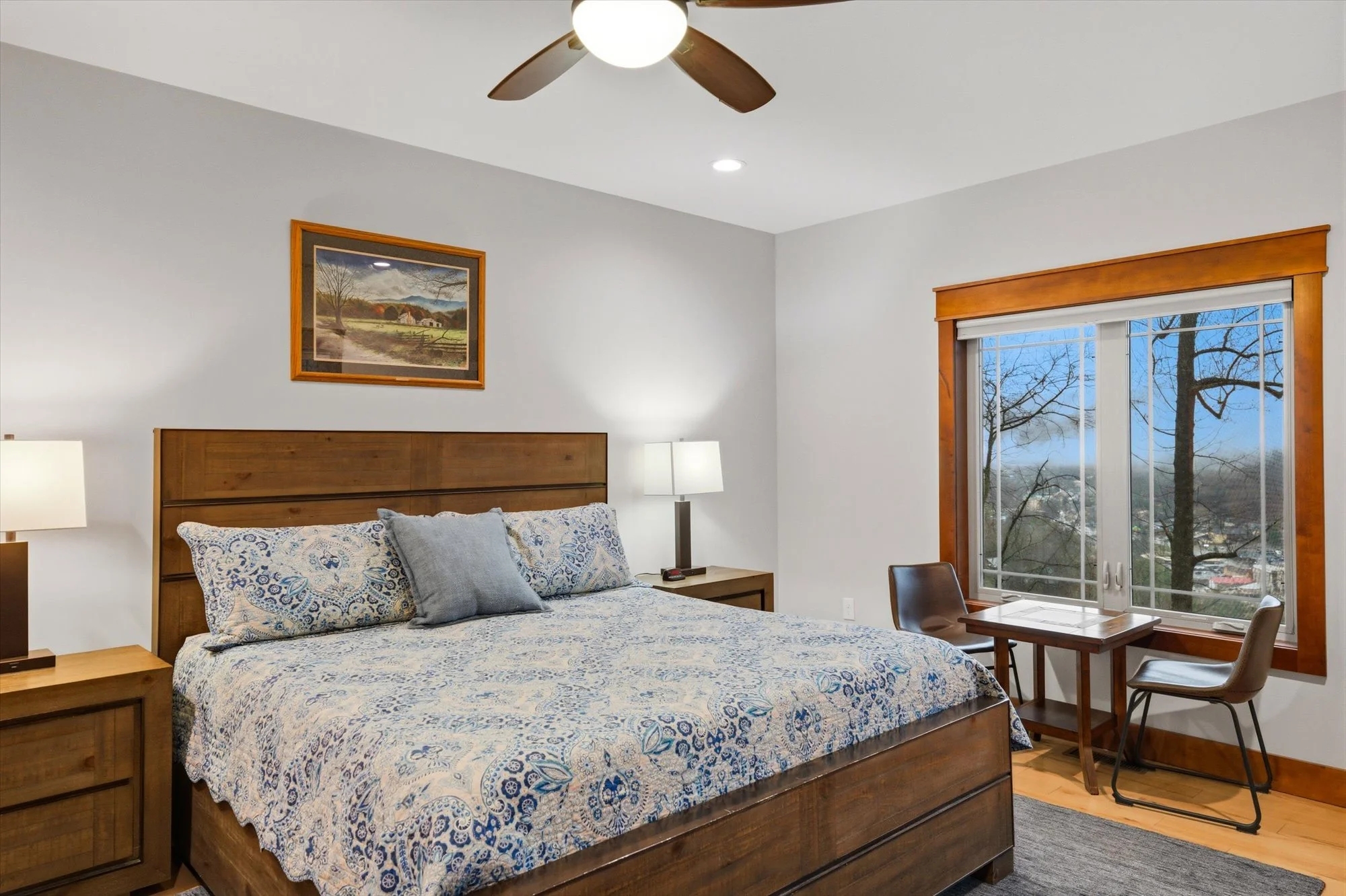 A cozy bedroom featuring a large wooden bed with a blue and white patterned bedspread and pillows. There are two wooden nightstands with lamps on either side of the bed. A framed landscape painting hangs above the bed. A window with a wooden frame sh
