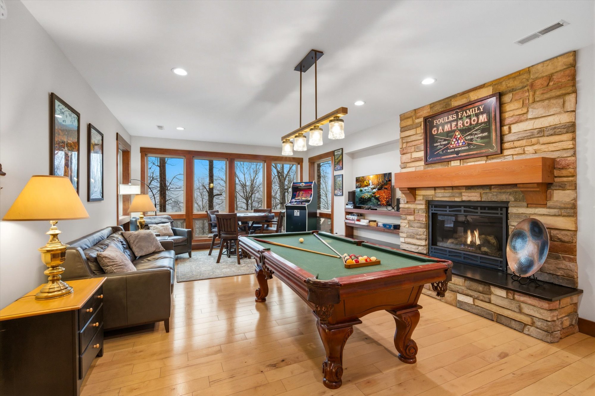 Living room with a pool table, fireplace, TV, and seating area, large windows showing trees outside.