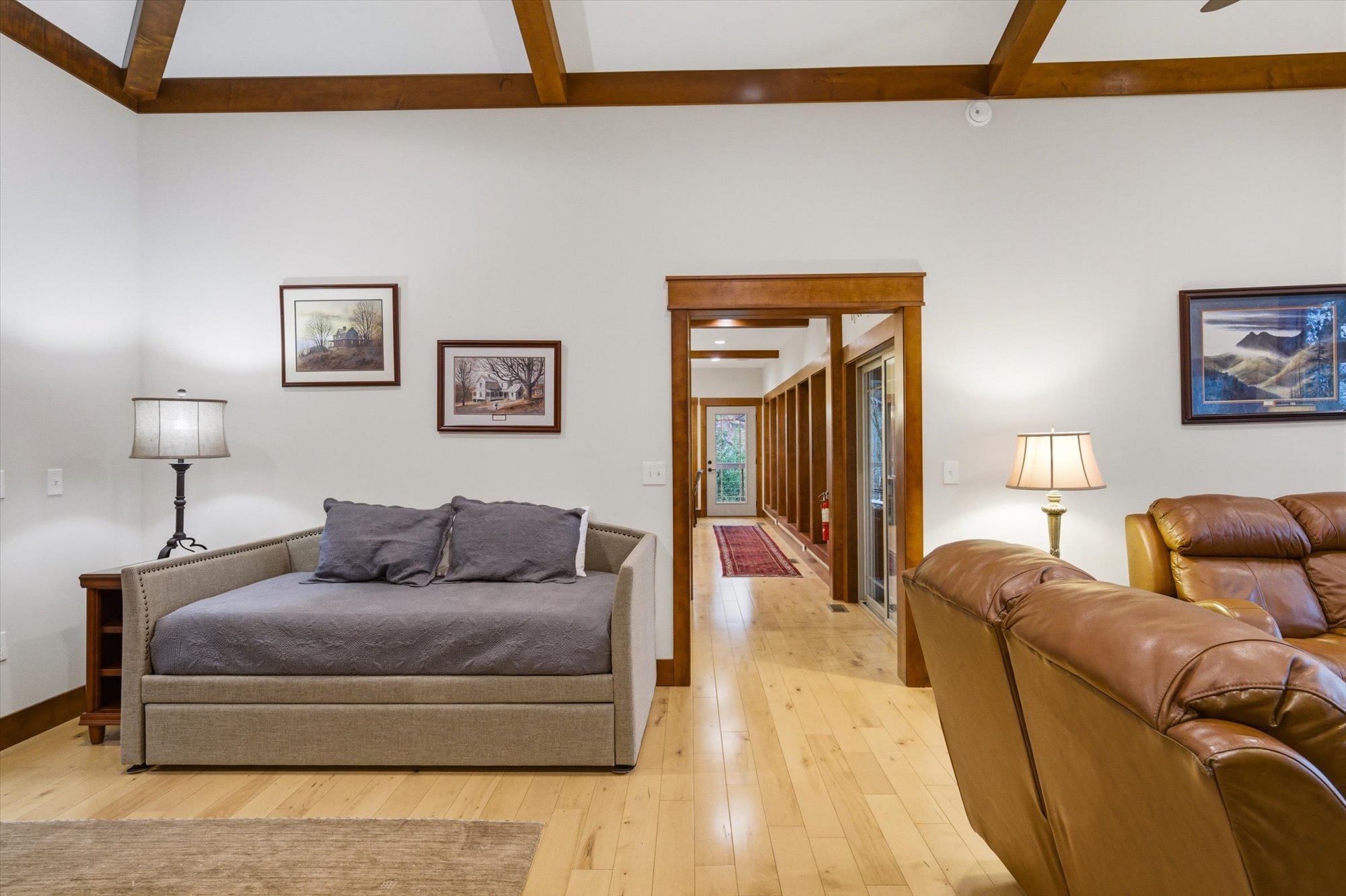 Living room with wooden floors, white walls, and wooden beams on the ceiling. Furniture includes a grey daybed with pillows, a brown leather sofa, and two lamps with beige shades. Framed landscape pictures hang on the walls. A hallway with a rug and 
