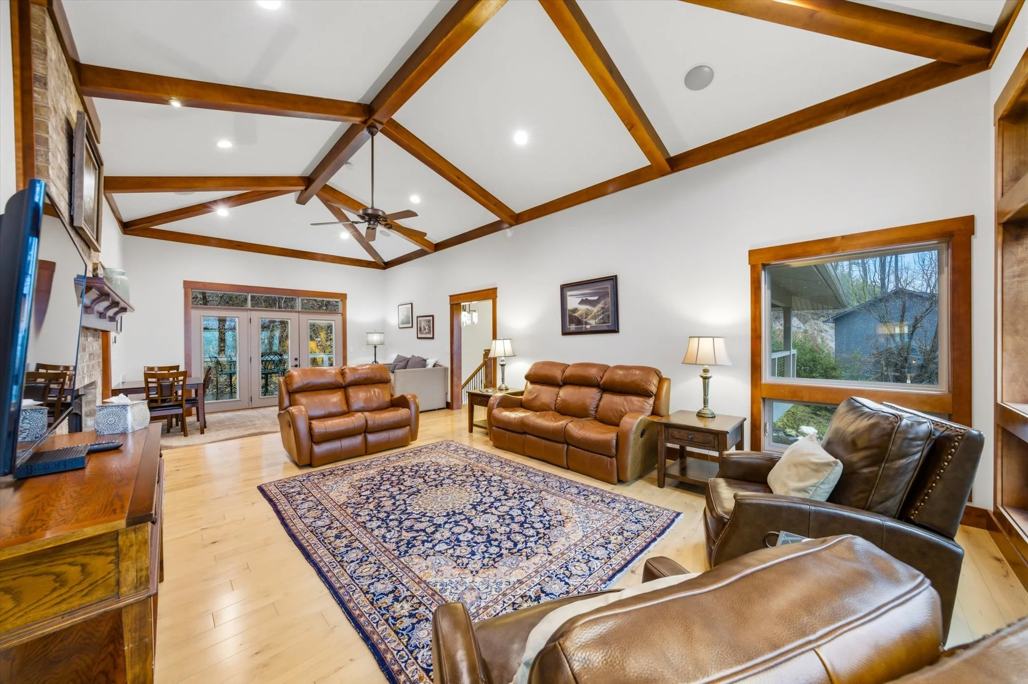 Living room with vaulted ceiling, wooden beams, hardwood flooring, and large windows showing outdoor trees. Contains leather sofas, armchair, area rug, side tables, lamps, and a fireplace with a TV above.