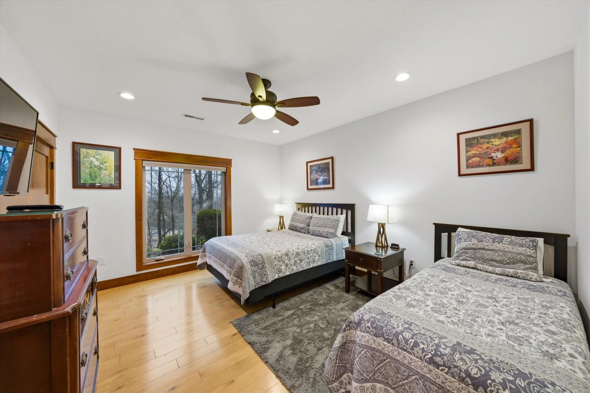 Two beds in a bedroom with hardwood floors, white walls, a ceiling fan, a window with a view of trees, and wooden trim. The beds have patterned bedspreads and pillows, with lamps on bedside tables and framed artwork on the walls.