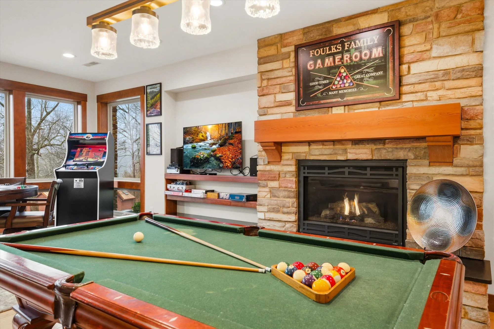 A cozy game room with a pool table, an arcade game, a television showing nature scenery, and a brick fireplace with a wooden mantel and a television above it. Large windows let in natural light, and wall art decorates the space.