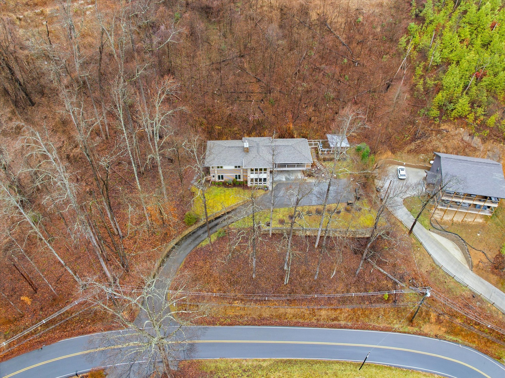 Aerial view of a residential area with multiple houses surrounded by leafless trees and a winding road. One house has a driveway with a parked car, and the area appears to be in late autumn or winter.