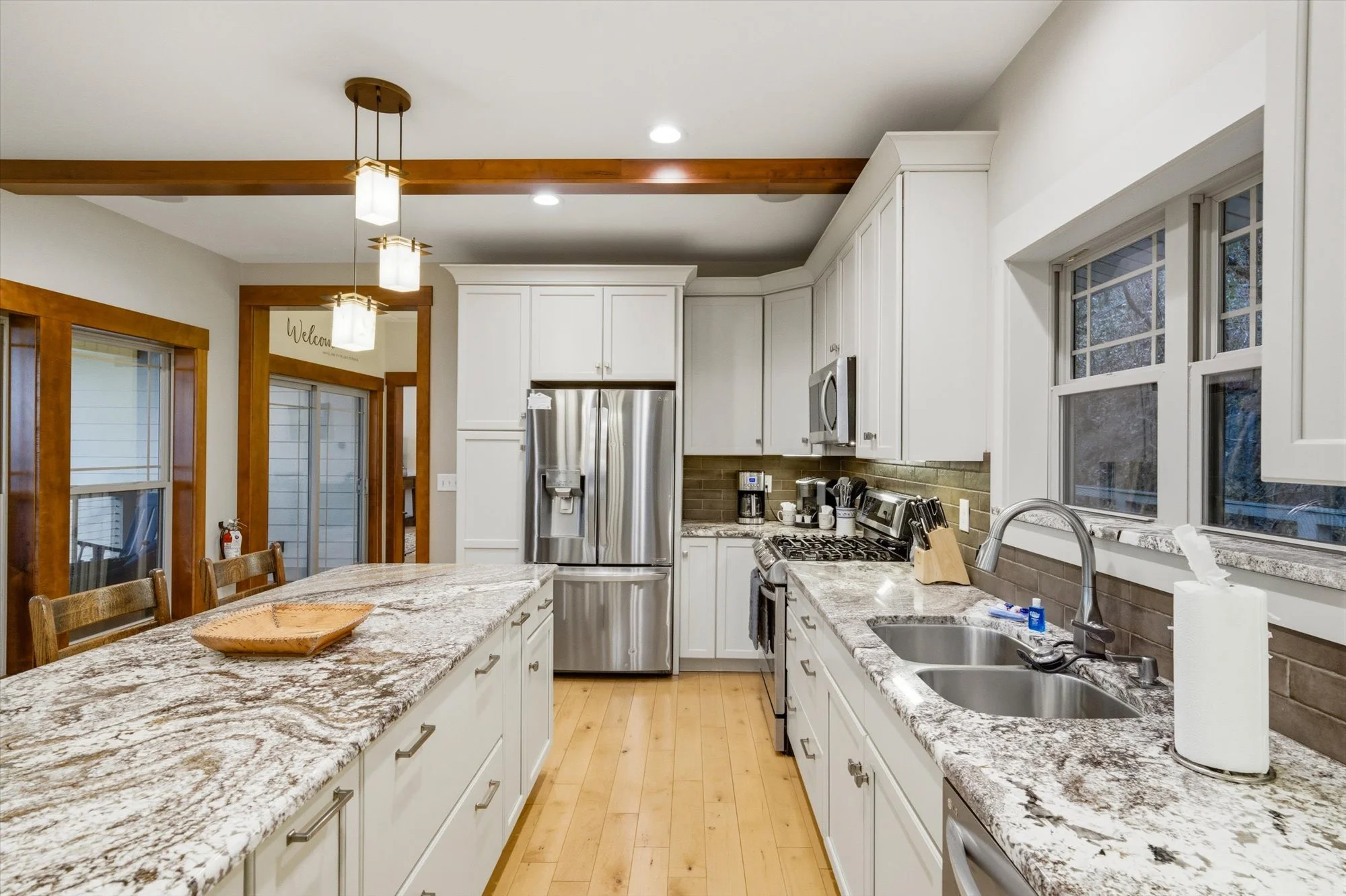 Kitchen with white cabinets, granite countertops, stainless steel refrigerator, stove, microwave, and a double sink. Wooden trim around the windows and door, hardwood flooring, and modern ceiling lights.