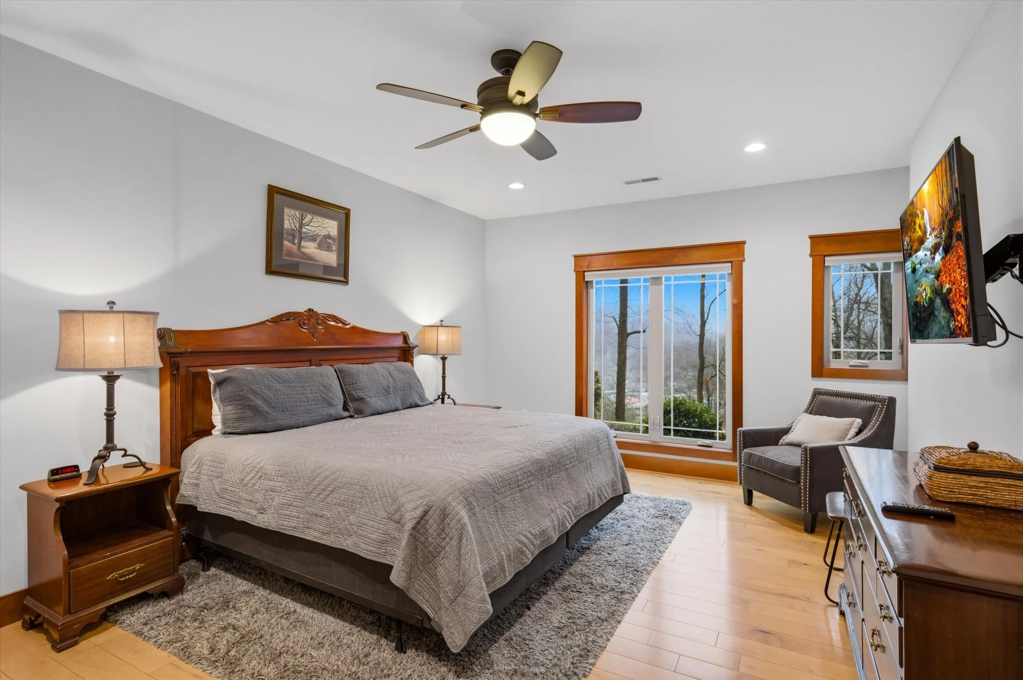 A bedroom with a wooden bed frame, gray bedding, two nightstands with lamps, a gray armchair with a pillow, a dresser with a basket, a wall-mounted TV, and two windows showing trees outside.