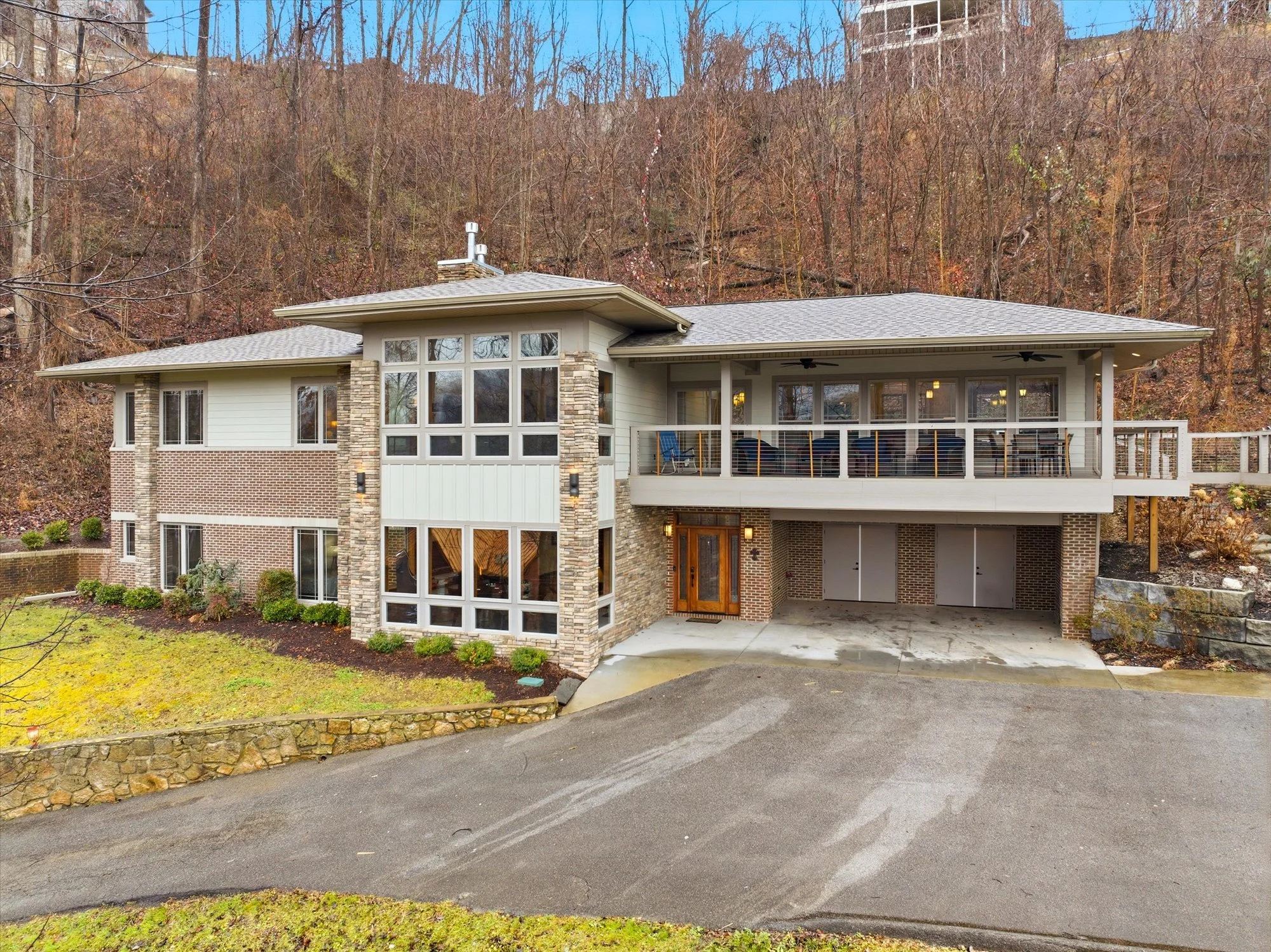 Modern multi-story house with large windows, a balcony, and a garage, situated on a hillside with wooded background and a stone wall in the front yard.