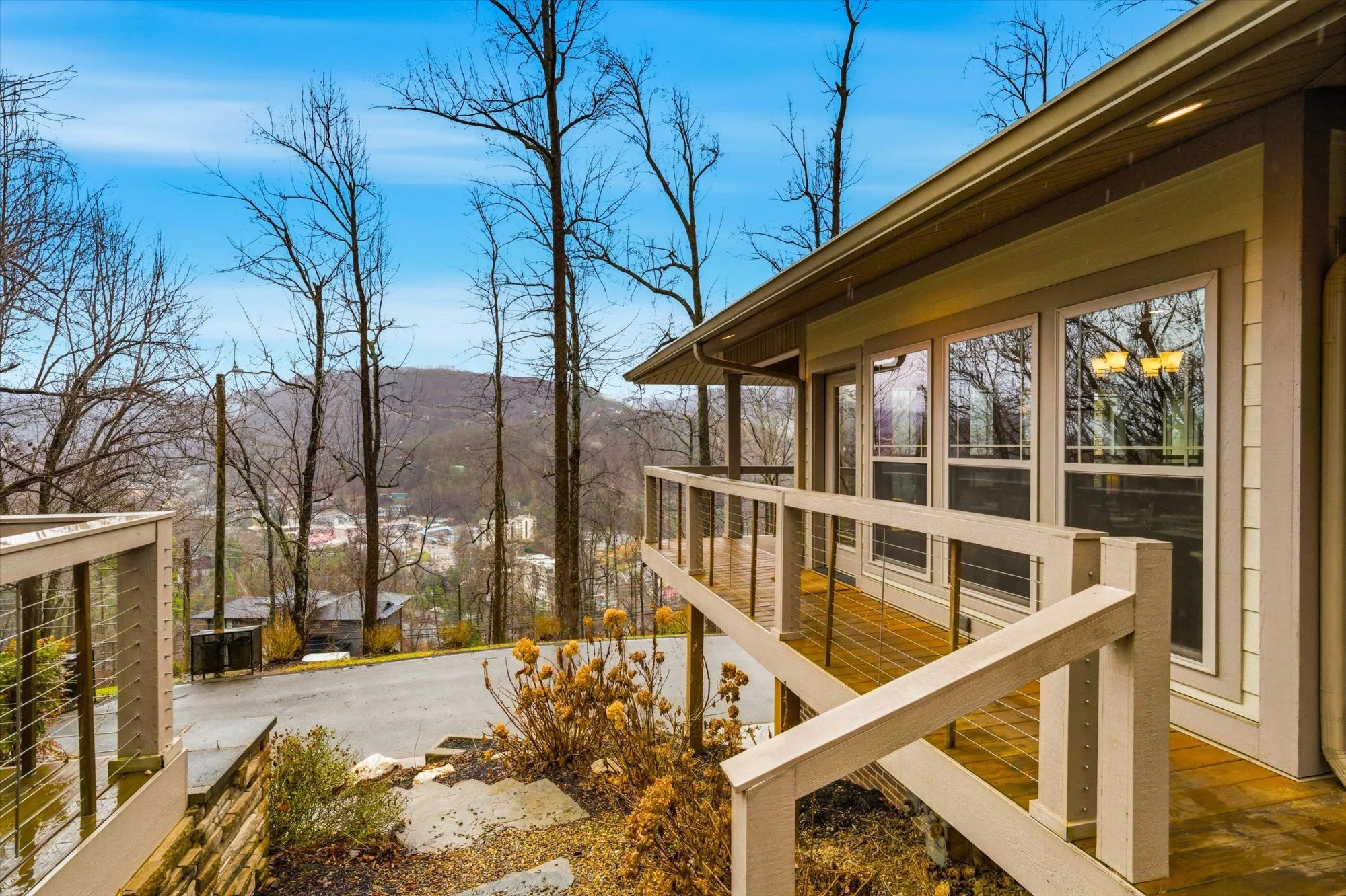 View from a house balcony with wooden railing, overlooking leafless trees, distant hills, and a small town in the background, during fall or winter.