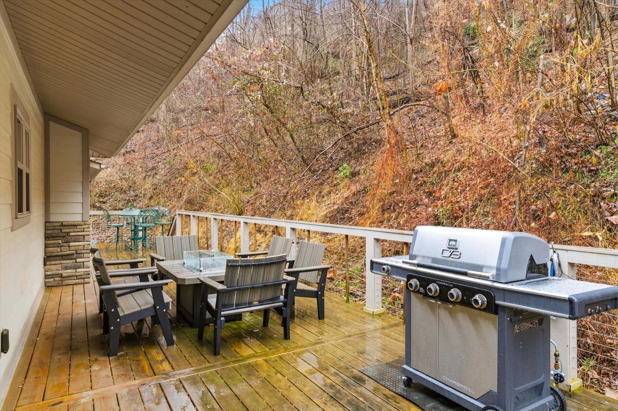 A wooden deck with outdoor furniture, including a dining table and chairs, and a gas grill, with a wooded hillside in the background.