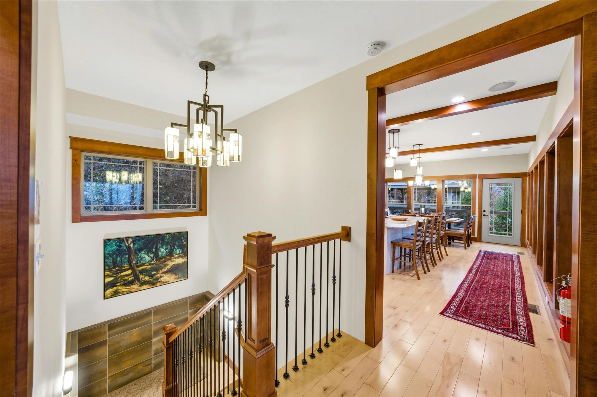 A view of an open interior space showing a staircase, a hallway, and a dining area with large windows and a glass door leading outside. The space features wooden accents, a modern chandelier, and a red rug on the wooden floor.