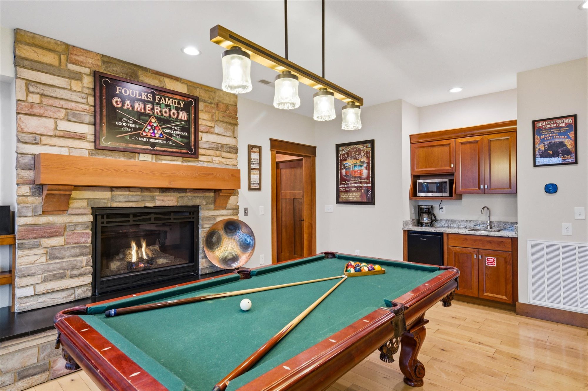 A basement game room featuring a pool table, a stone fireplace with a framed sign above it, a wooden cabinet with a microwave, a coffee maker, artwork on the walls, and a ceiling light fixture.