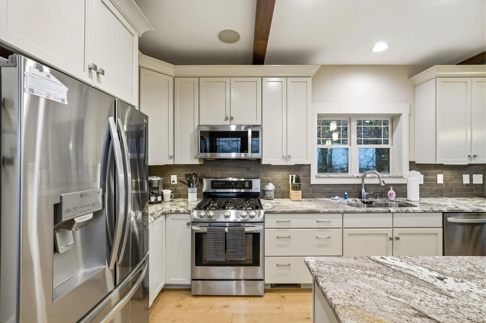Modern kitchen with white cabinets, stainless steel appliances, granite countertops, and a window above the sink showing outdoor trees.