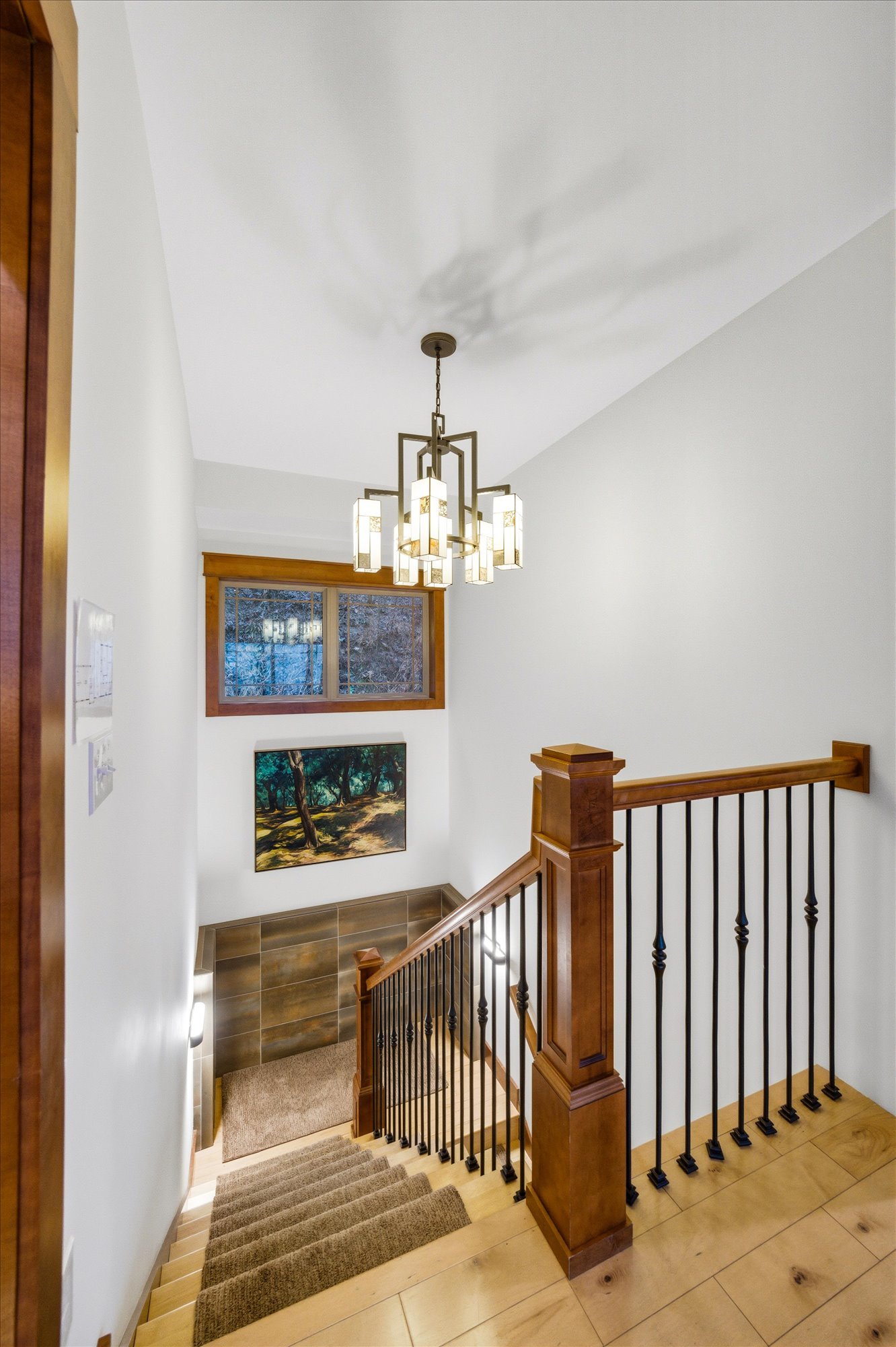 Interior view of a staircase with a wooden handrail and black metal balusters, leading down to a landing with wood and tile flooring, a painting of a forest scene on the wall, and a window with wood trim, illuminated by a modern chandelier hanging fr