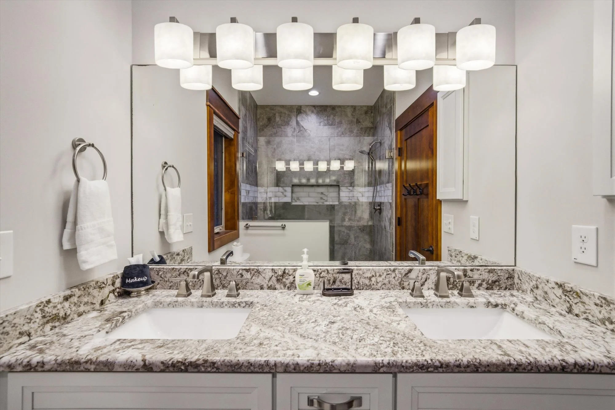 A spacious bathroom featuring a granite double vanity with two sinks, a large mirror, and a modern light fixture above. Behind the vanity is a walk-in shower with gray and white tile, a glass door, and a rainfall showerhead. The room has white walls,