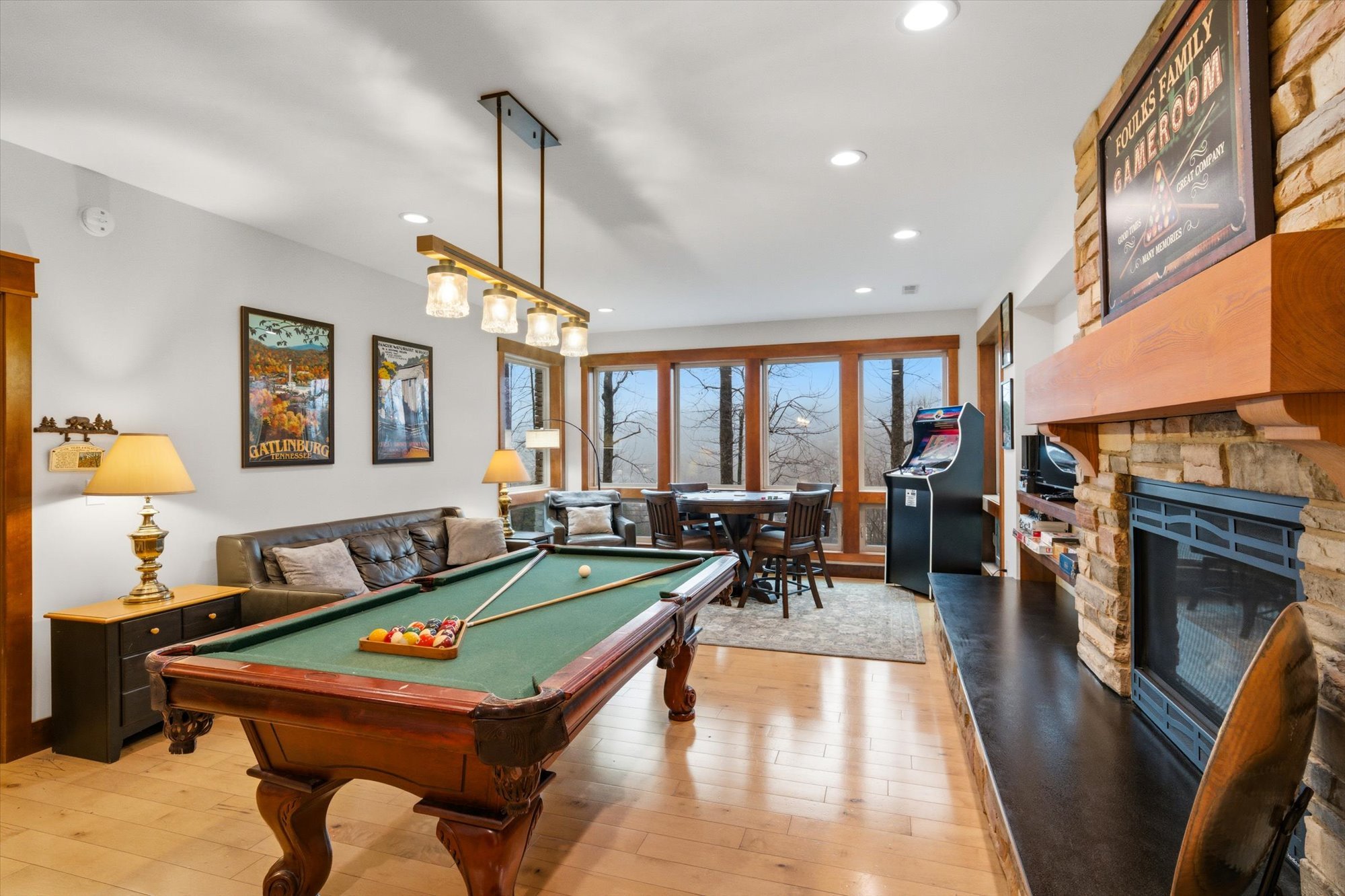 Living room with a pool table, a leather sofa, and a fireplace. There is a large window with a view of trees, a dining table with chairs, and an arcade game machine. The room has wooden floors and white walls decorated with framed posters.