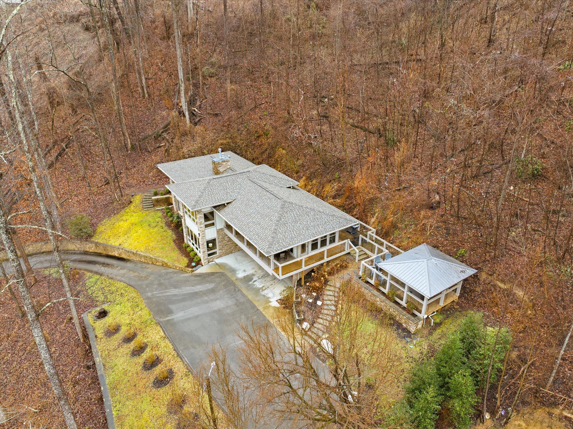Aerial view of a house with a gray roof, surrounded by brown trees and a driveway with a small circular plant.