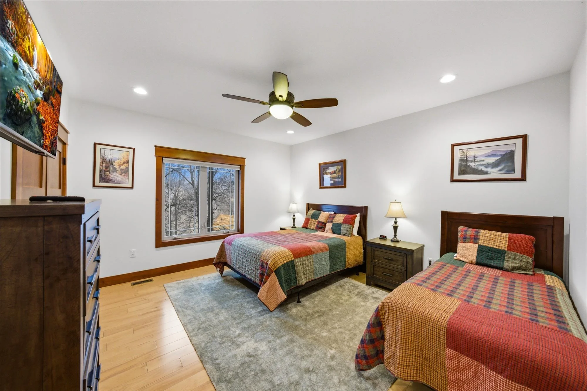 A bedroom with two beds covered in colorful patchwork quilts, a large window showing trees outside, and a ceiling fan with lights, wooden furniture, framed landscape paintings, and a gray area rug.