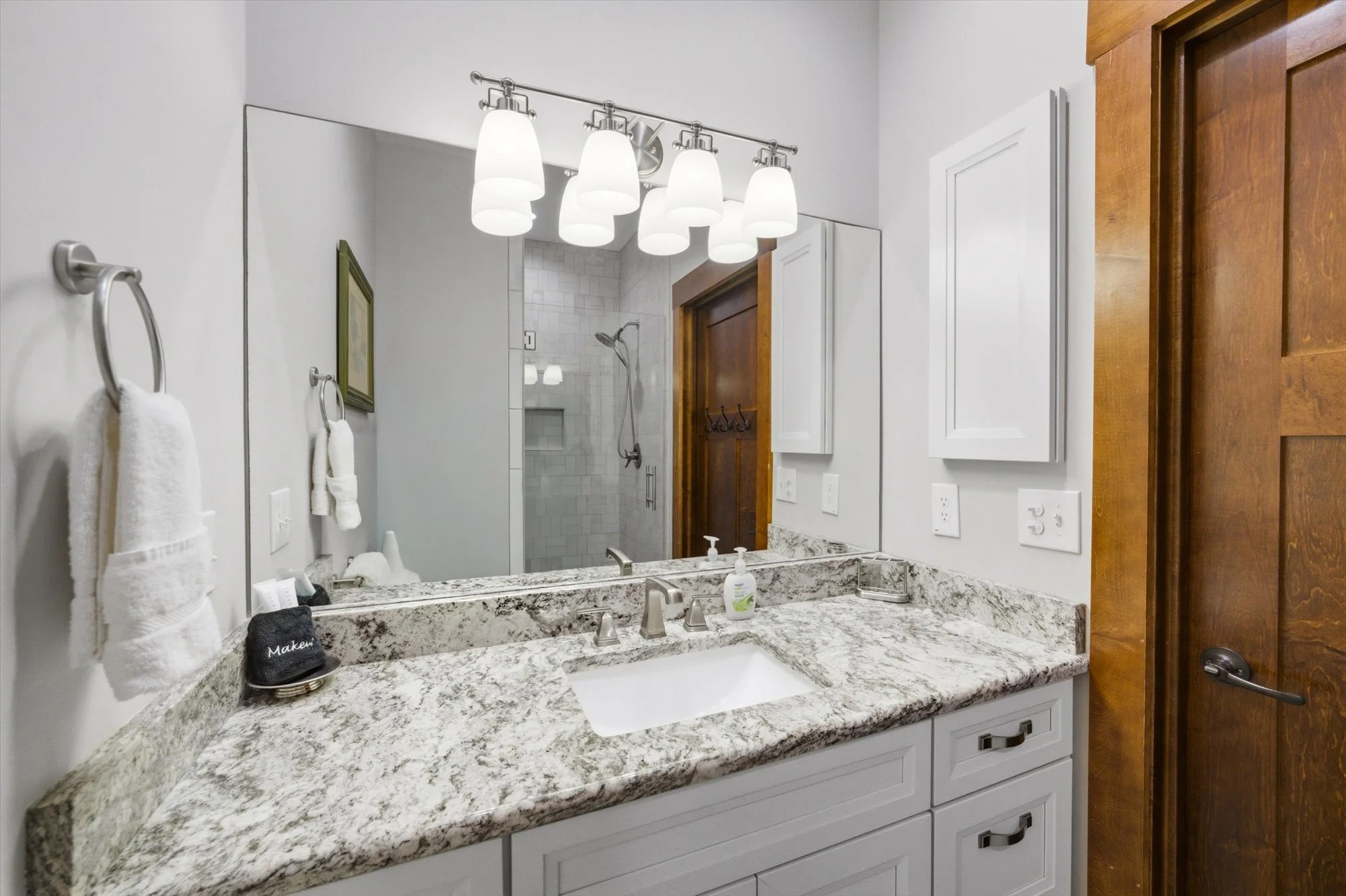 Bathroom vanity area with white cabinets, a granite countertop, a large mirror, and a four-light fixture above. A towel is hanging on a towel ring, and a soap dispenser is on the counter. A wooden door is on the right, and a separate shower with a gl