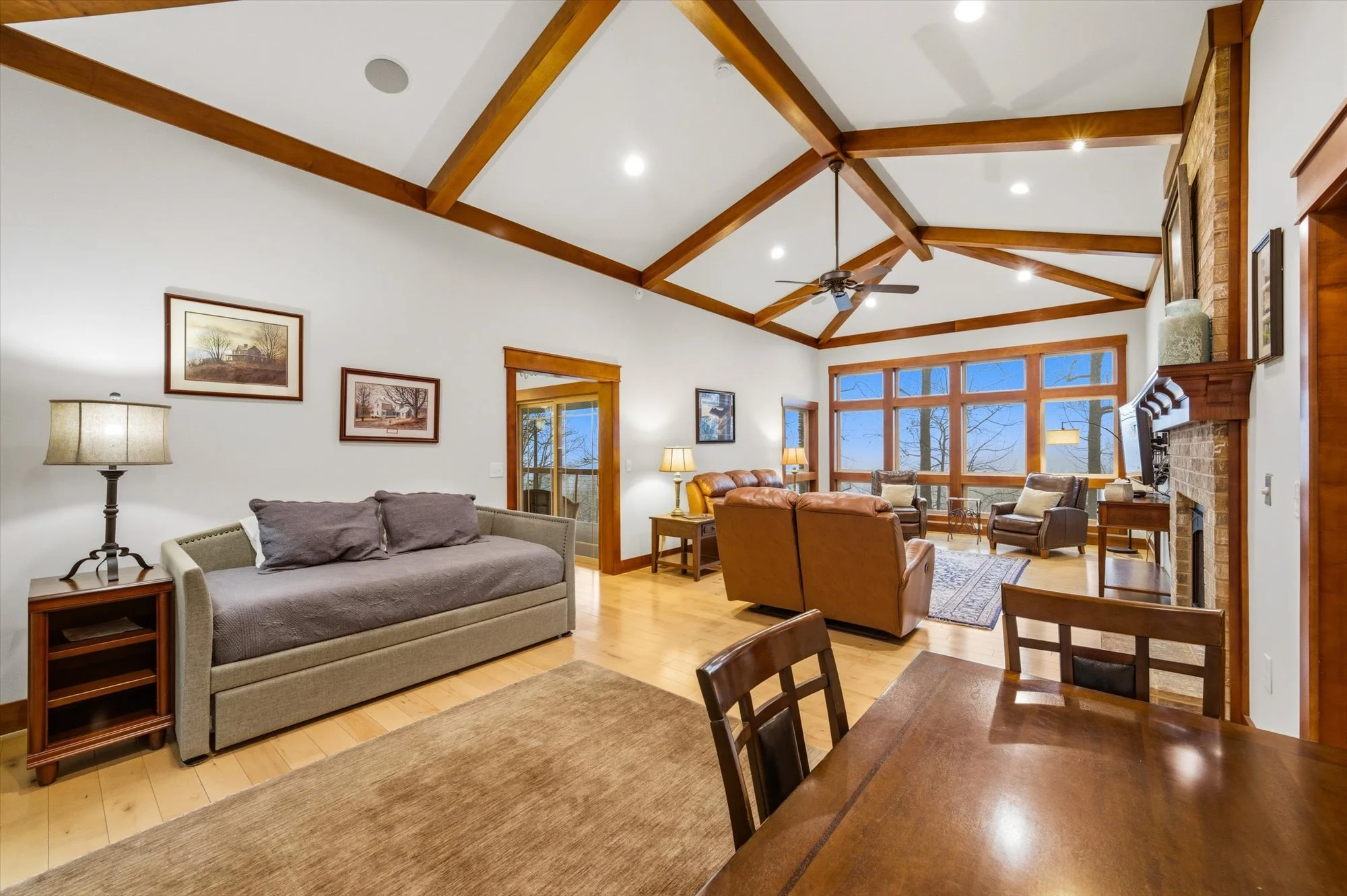 Living room with vaulted ceiling, exposed wooden beams, large windows, brick fireplace, and various chairs and sofas.