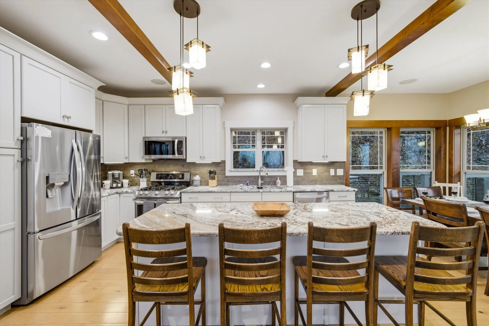 Kitchen with white cabinets, stainless steel appliances, granite island, wooden chairs, pendant and recessed lighting, and large windows.