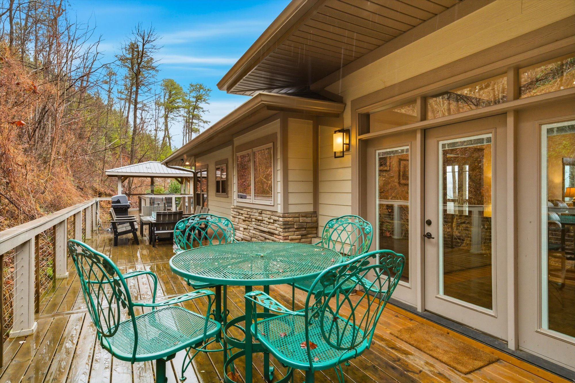 A wooden deck with outdoor furniture, including a green metal table and chairs, a grilling station, and a covered seating area, attached to a house with large windows and a sliding glass door, surrounded by trees.