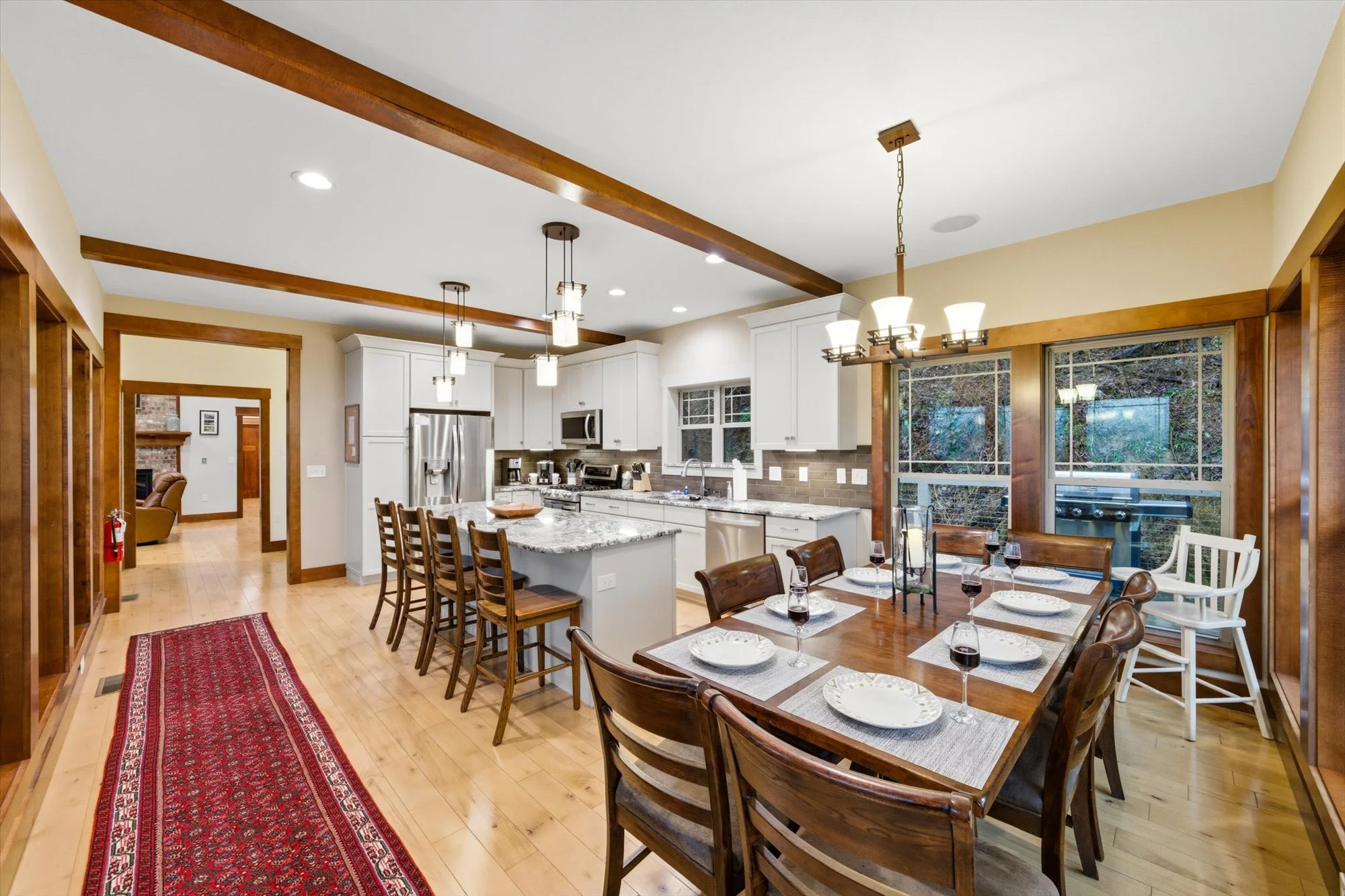 A spacious kitchen and dining area with white cabinets, stainless steel appliances, and a large wooden dining table set for six, with wine glasses and plates, in a home with hardwood floors and large windows.
