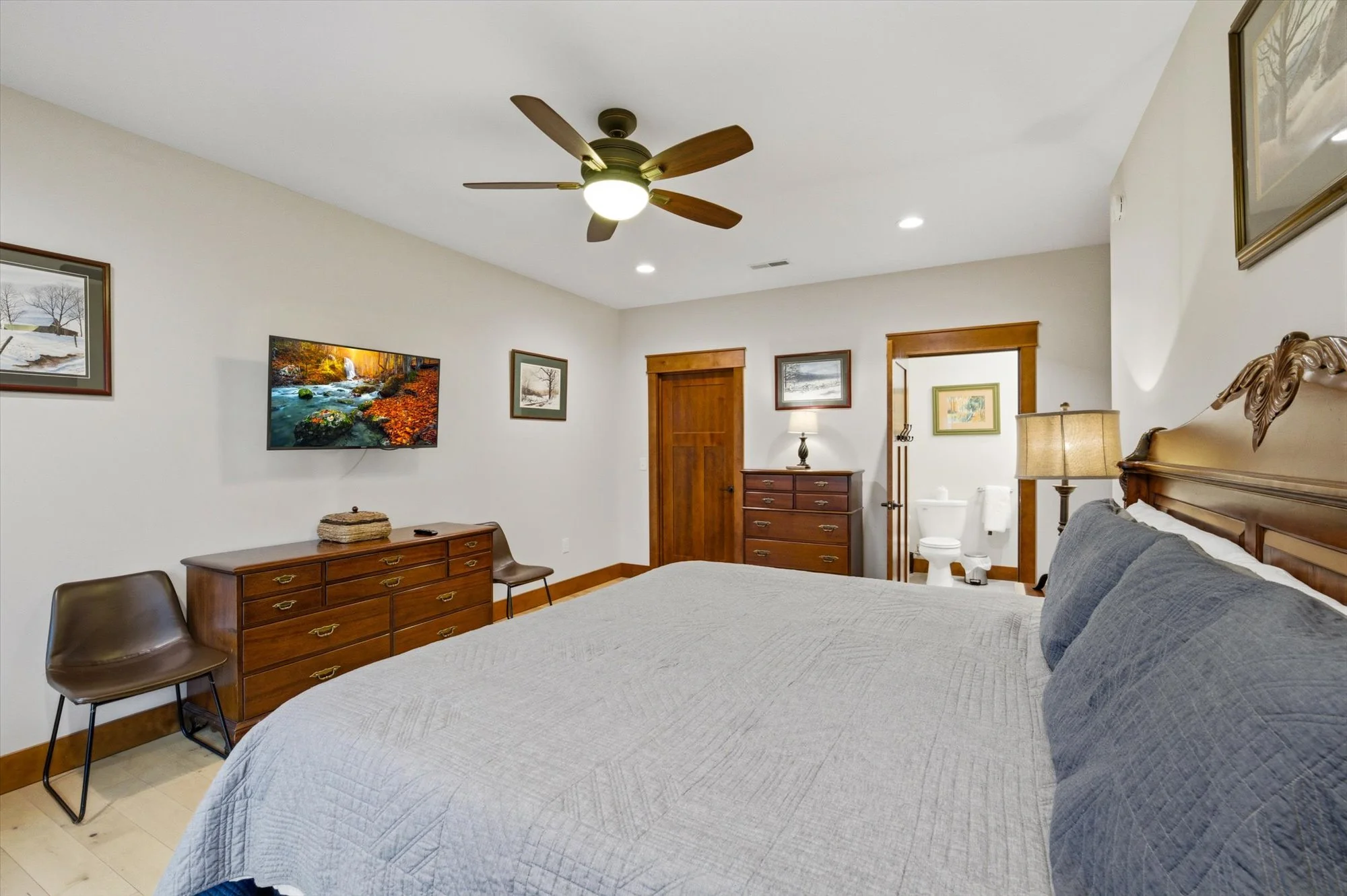 A bedroom with a large wooden bed, nightstand, and dresser, decorated in neutral tones with paintings on the walls, a ceiling fan, and an en suite bathroom.
