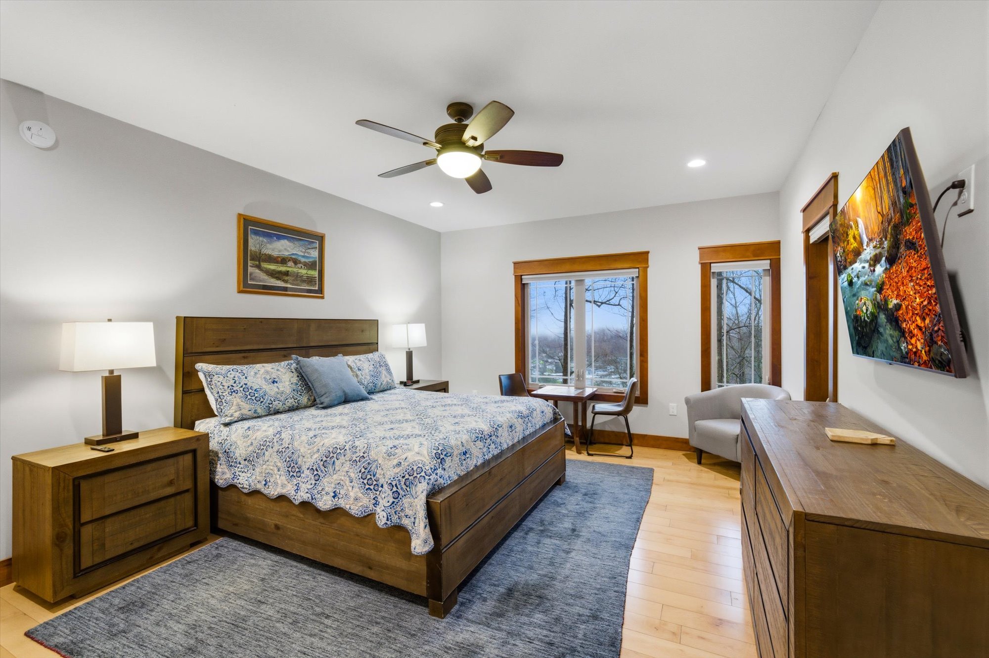 Modern bedroom with a king-size bed, wooden furniture, two wall-mounted lamps, a ceiling fan, a small sitting area near large windows, a wall-mounted TV, and a landscape painting.