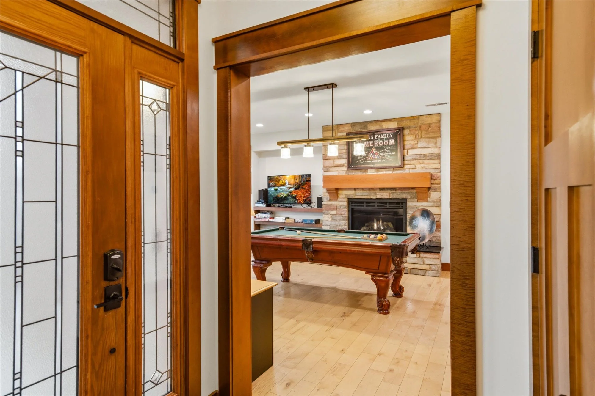 View of a game room with a pool table, brick fireplace, TV, and decorative wall signs, seen through a wooden doorway with stained glass panels.